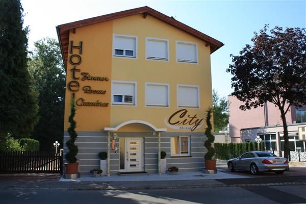 Žltá budova s nápisom "City Hotel", obklopená stromami a zaparkovaným autom.