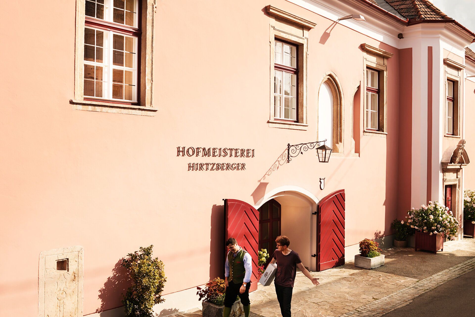 Die sanften Farben der Fassade und die einladenden Türen der Hofmeisterei strahlen eine herzliche Atmosphäre aus. Umgeben von blühenden Pflanzen und dem Charme der Wachau, lädt dieser Ort dazu ein, die traditionelle Wirtshauskultur Niederösterreichs zu erleben.