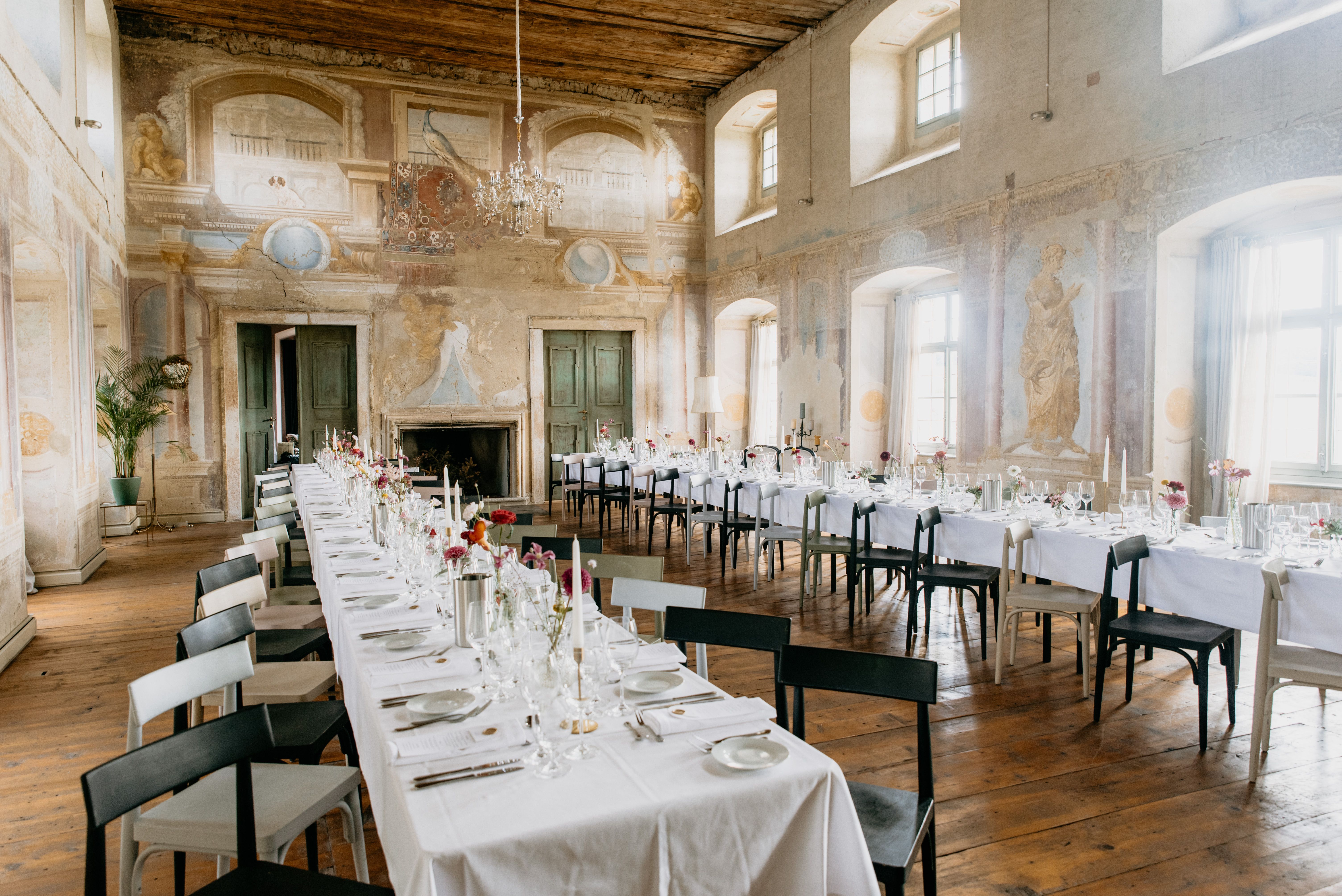 Elegantná banketová sála s dlhými prestretými stolmi, starožitnými nástennými maľbami a lustrami.