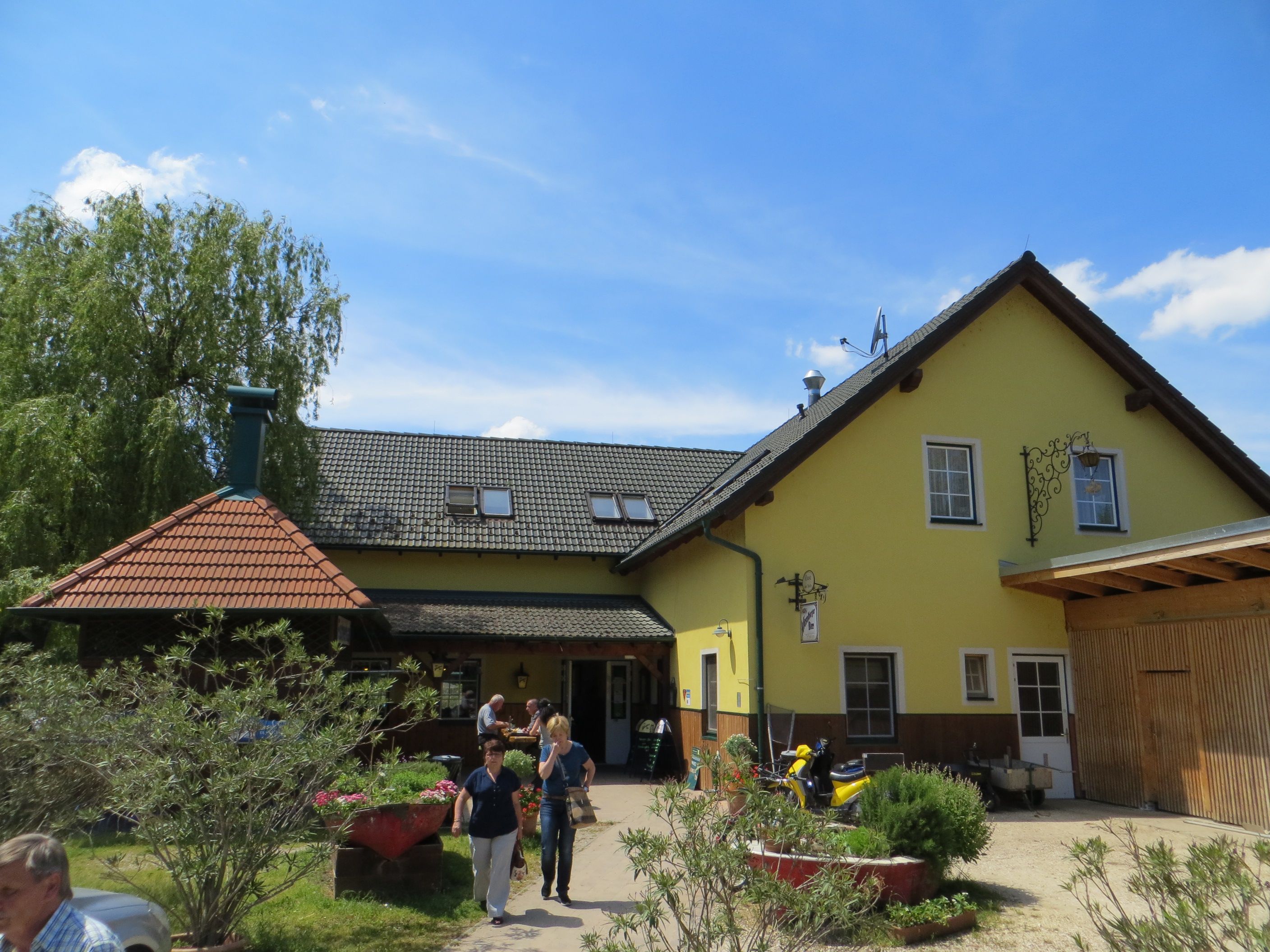 Žltý hostinec so sivou strechou a záhradou v popredí.