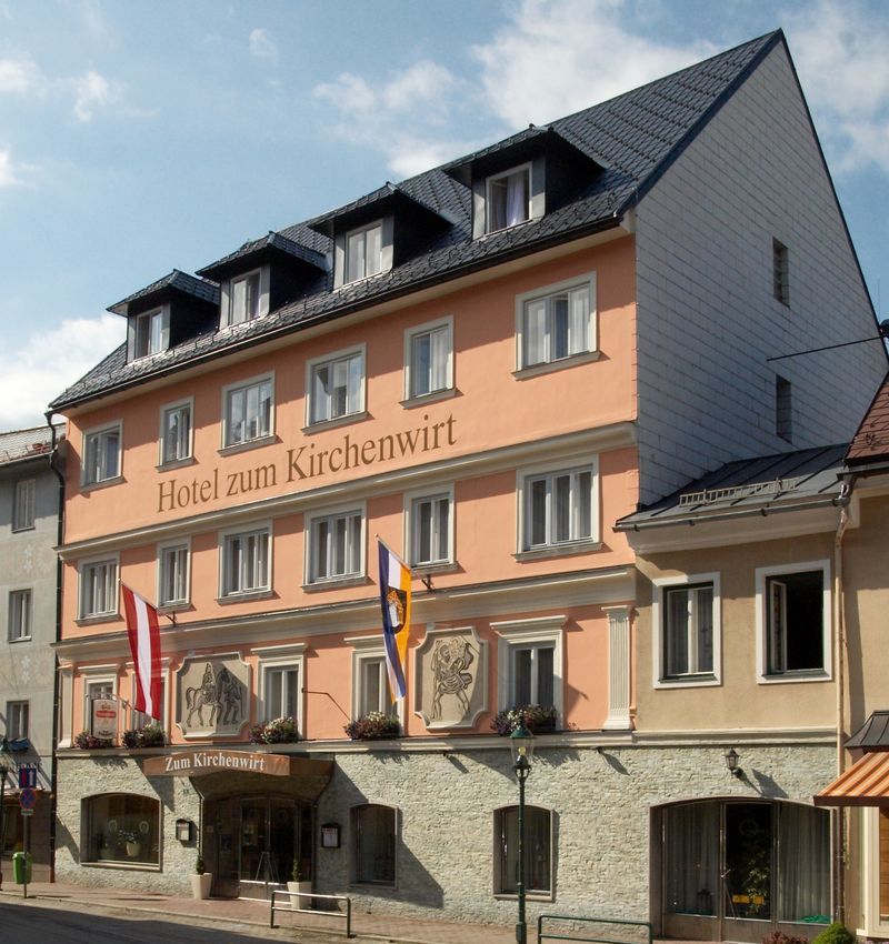 Fasáda hotela zum Kirchenwirt s vlajkami a modrou oblohou.