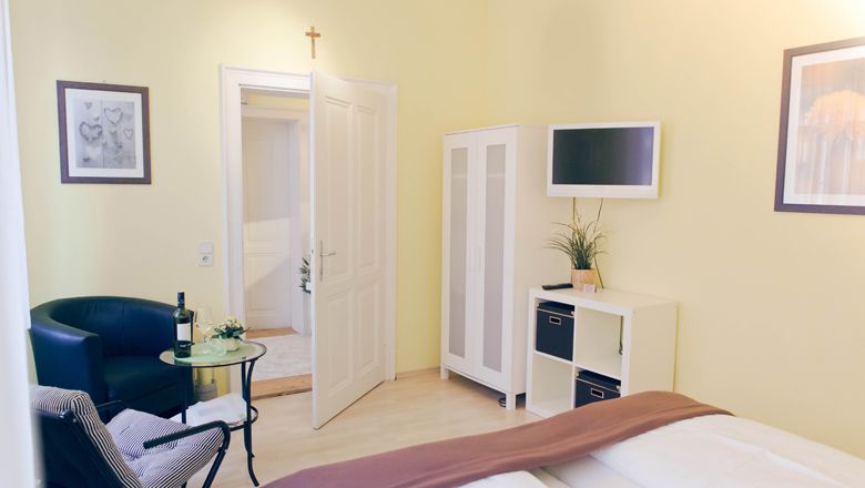 Svetlá izba pre hostí s posteľou, kreslom, stolom, šatníkovou skriňou a TV.