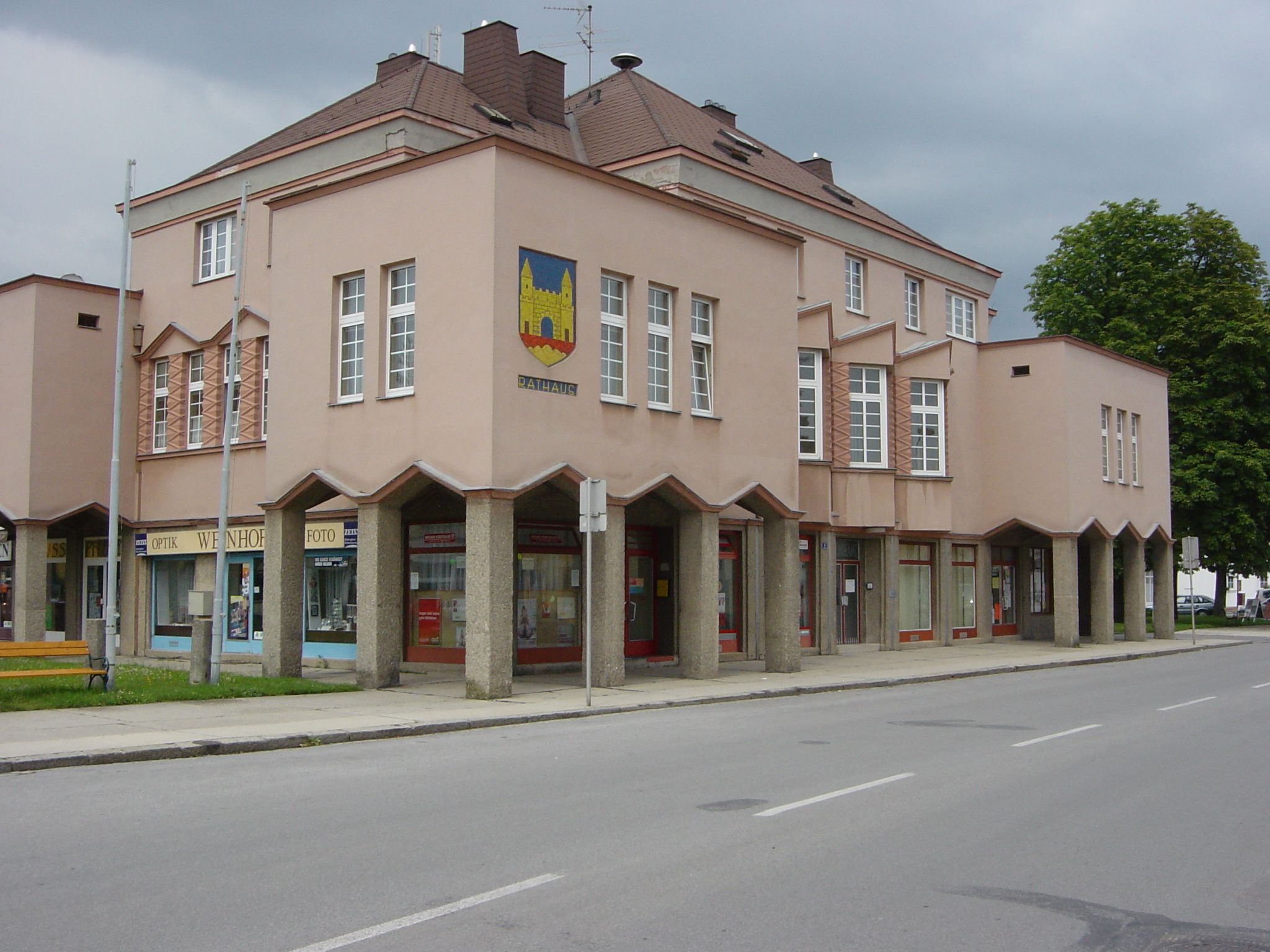 Moderná budova radnice s ružovou fasádou a veľkými oknami. Na nároží má erb a na prízemí obchody.
