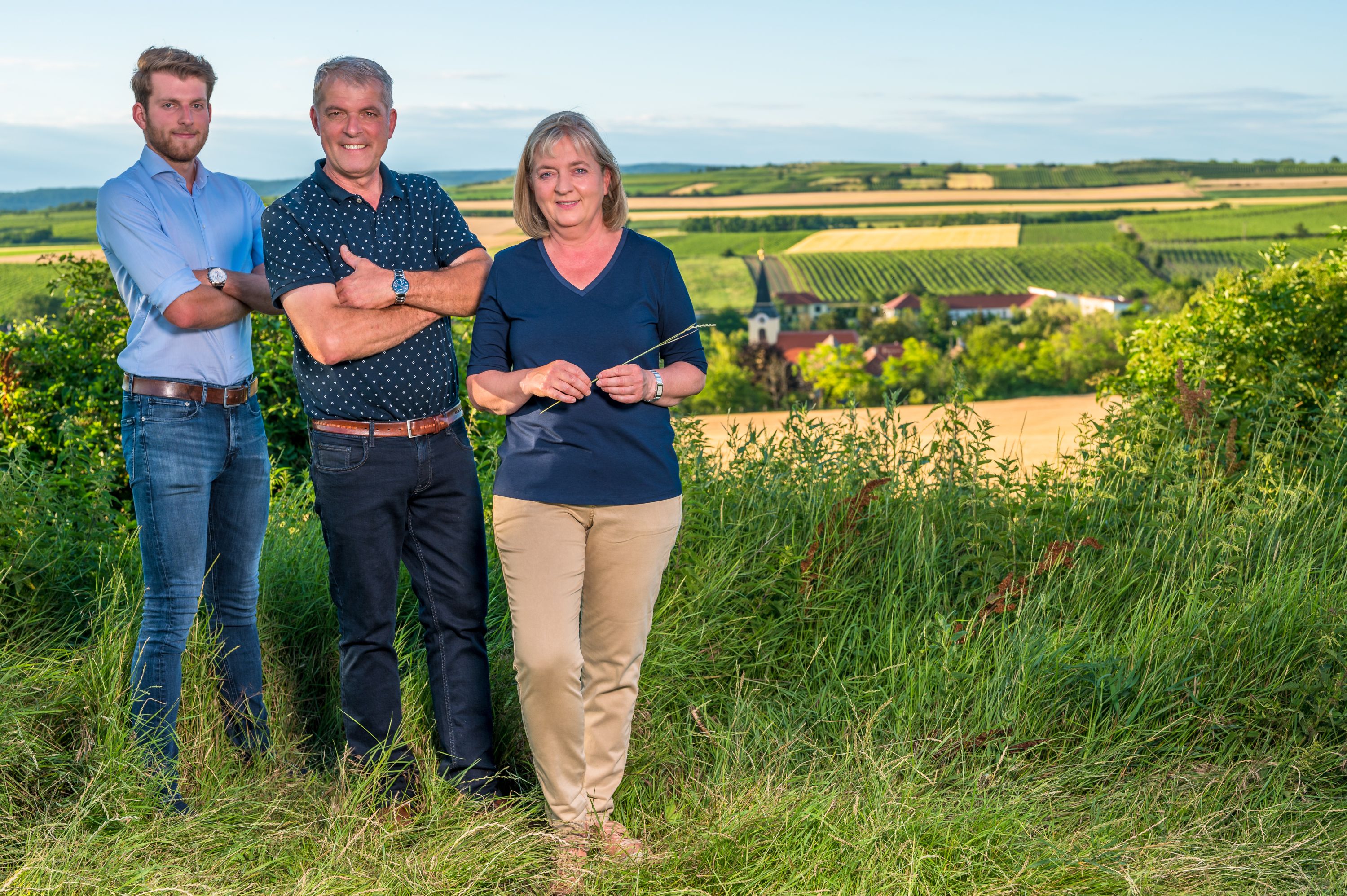 Traja ľudia stoja vo vinici s výhľadom na vidiecku krajinu v pozadí.