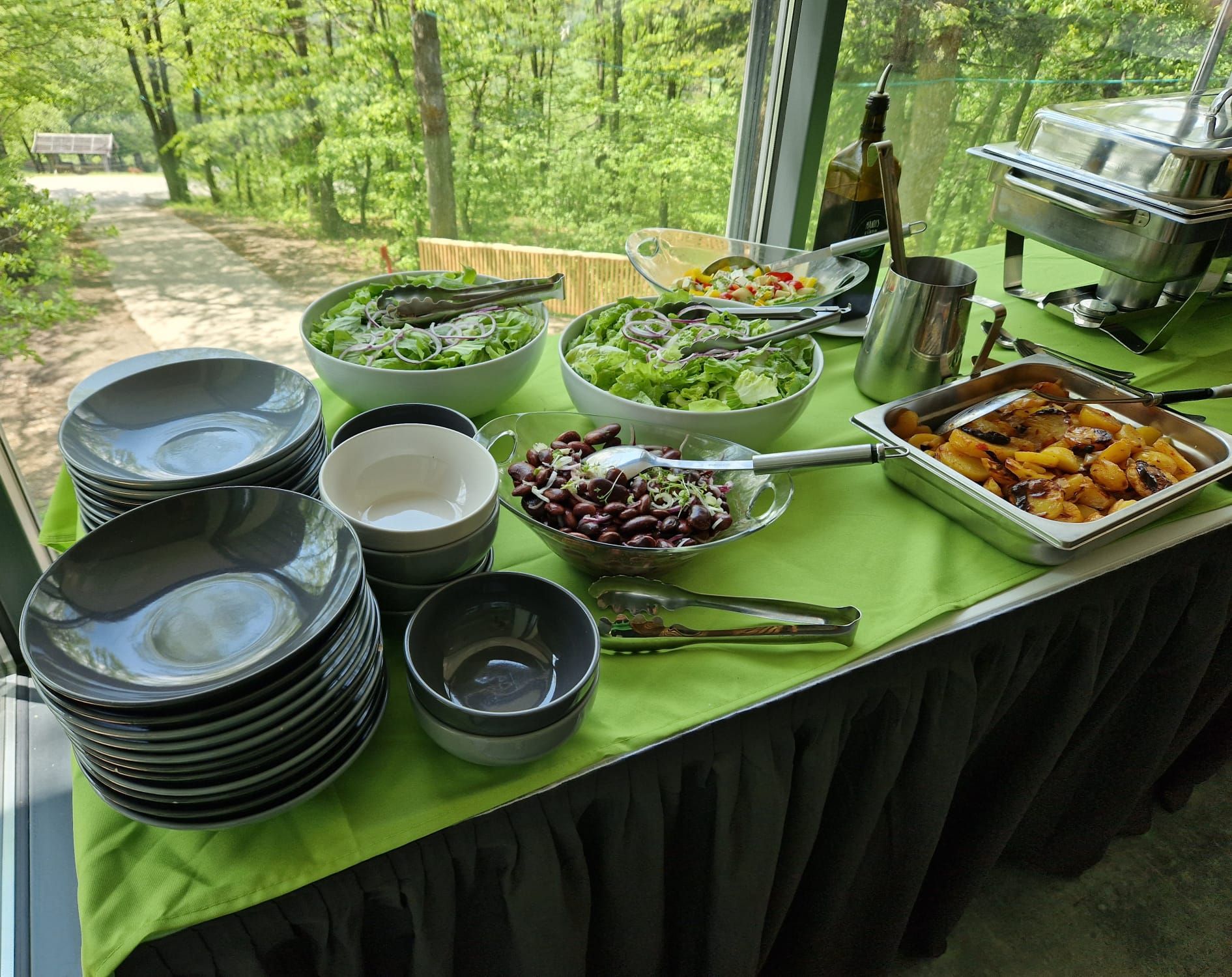 Vonkajší bufet s rôznymi šalátmi, taniermi, zemiakmi a príbormi na zelenom obruse.