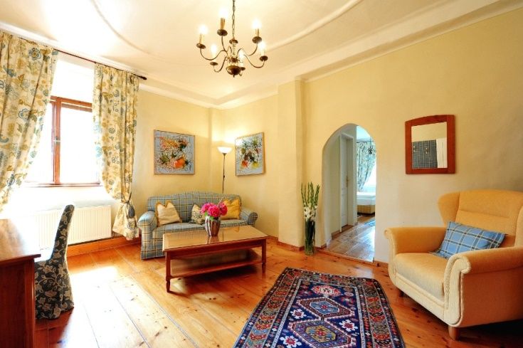 Útulná obývacia izba s drevenou podlahou, pohovkou, kreslom, kobercom a kvetmi.