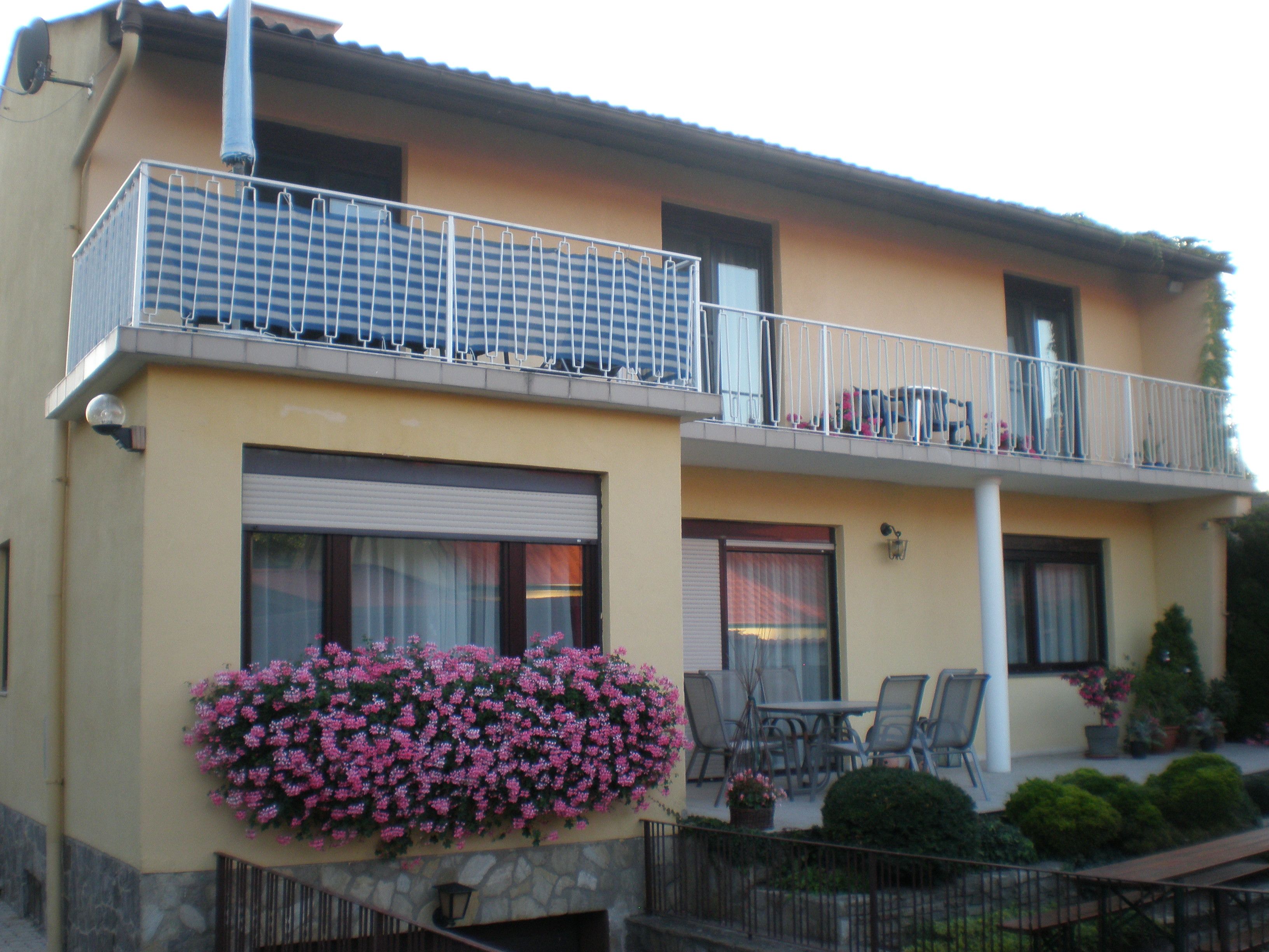 Dvojpodlažný dom s balkónom, kvetinami a záhradným nábytkom.