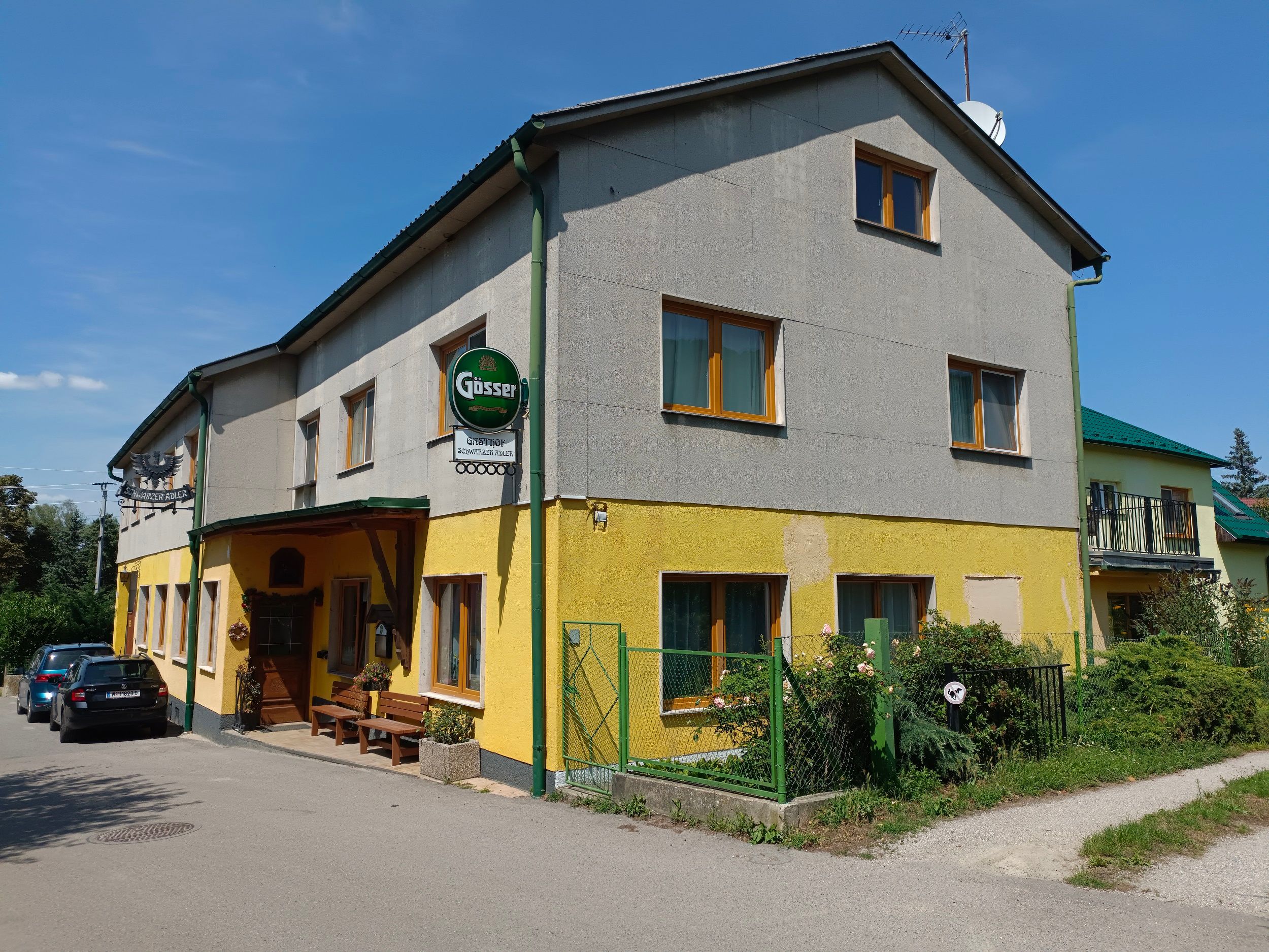 Pohľad zvonku na žltošedý hostinec s nápisom Gösser a zaparkovanými autami.