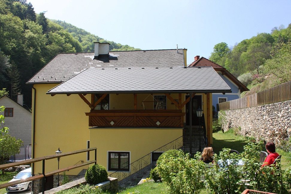 Žltý dom s balkónom, obklopený zelenou krajinou a kamenným múrom.