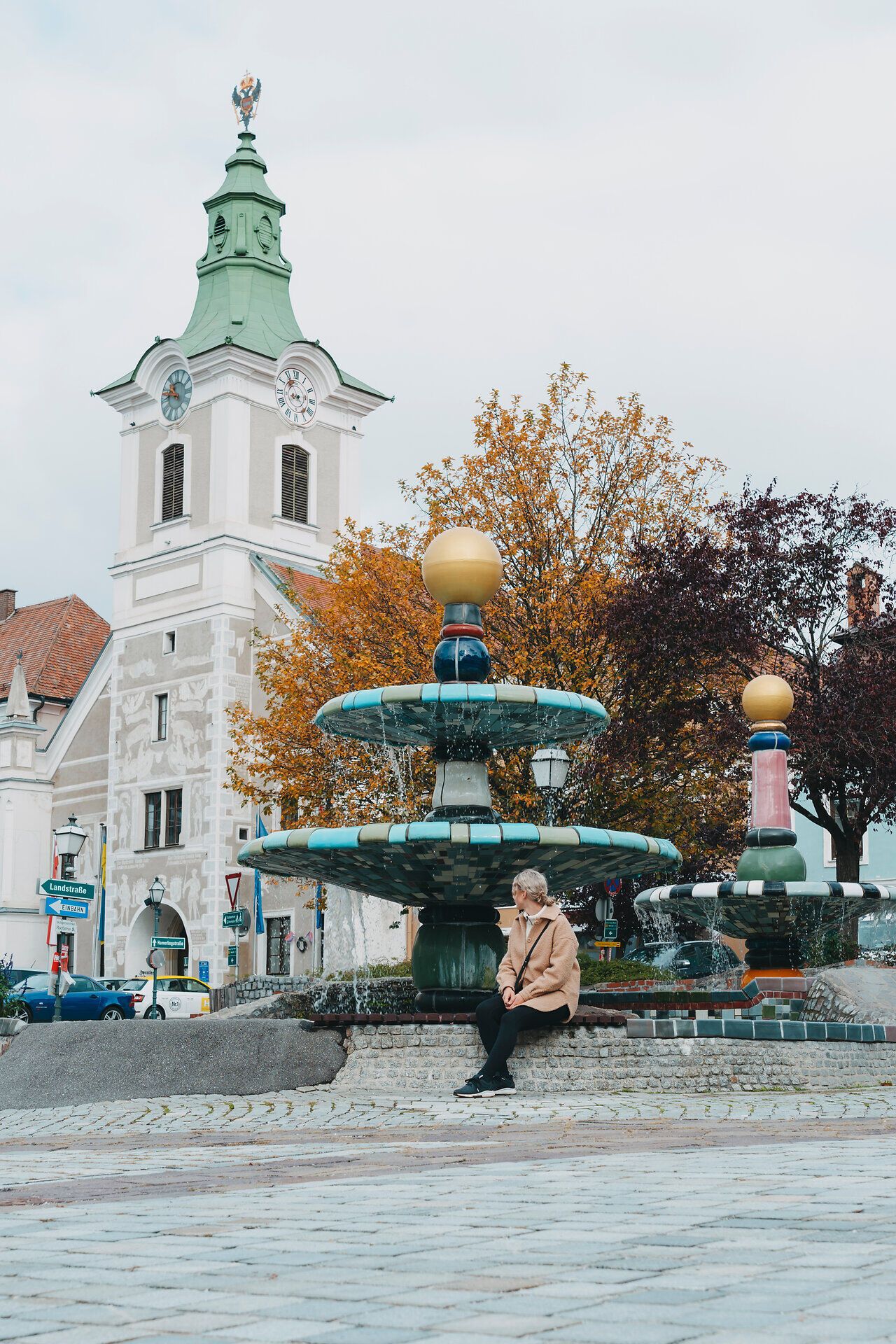 Der Herbst hüllt den Platz in sanfte Farben, während das Wasser des kunstvollen Brunnens sanft plätschert. Eine ruhige Atmosphäre lädt dazu ein, die Schönheit der Umgebung zu genießen und die kreative Architektur zu bewundern.