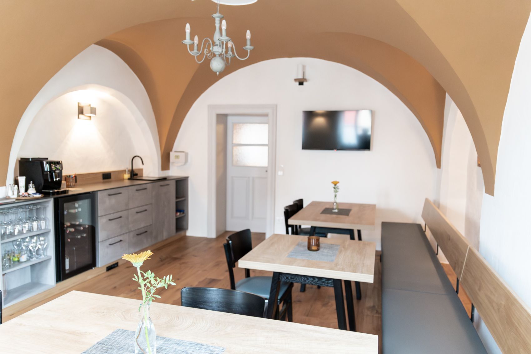 Útulná raňajková miestnosť s drevenými stolmi, stoličkami a lavicou. Na stene visí televízor a k dispozícii je kuchynka s kávovarom.