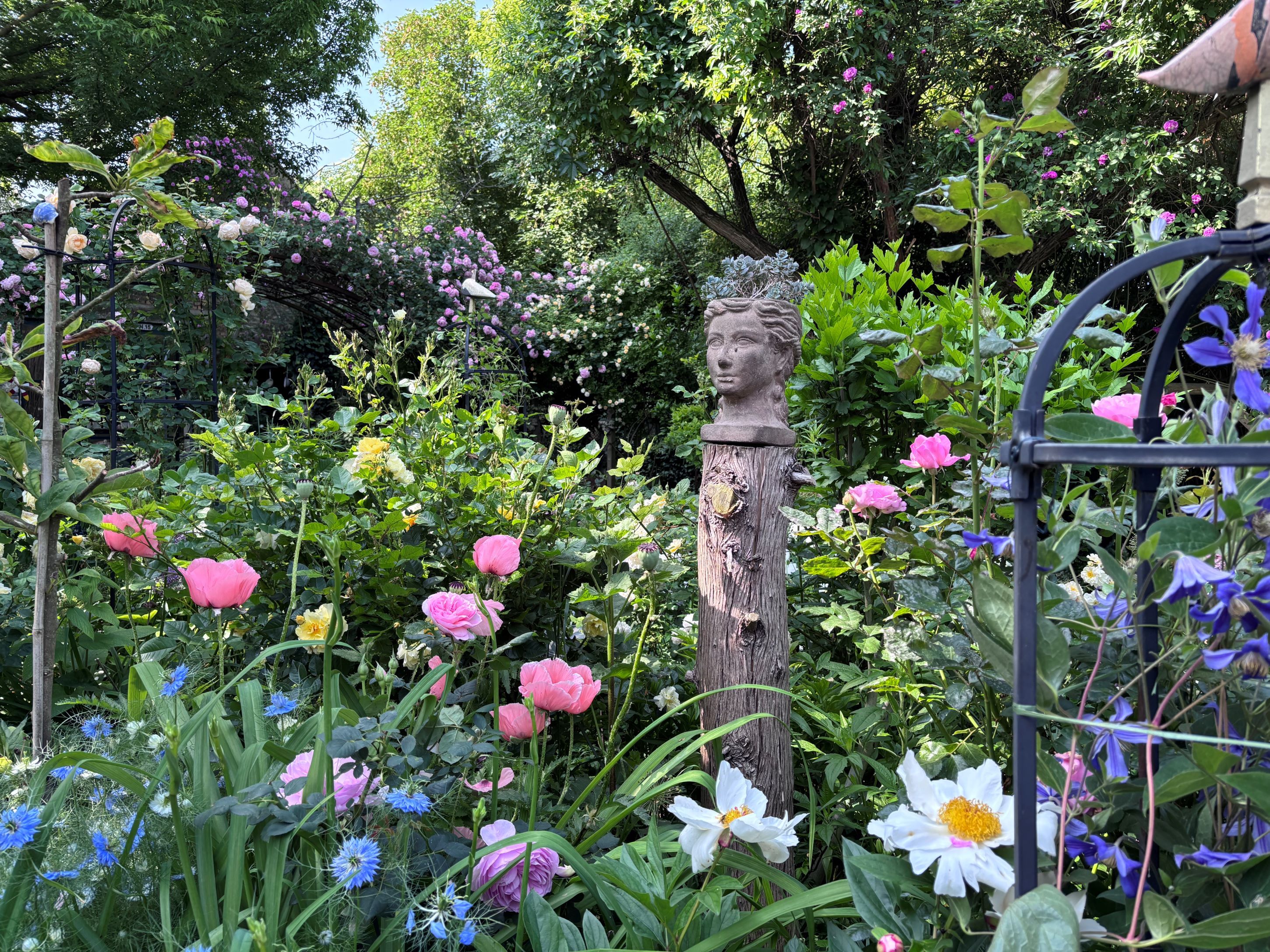 Rozkvitnutá záhrada s farebnými kvetmi a bustou na drevenom stĺpe.