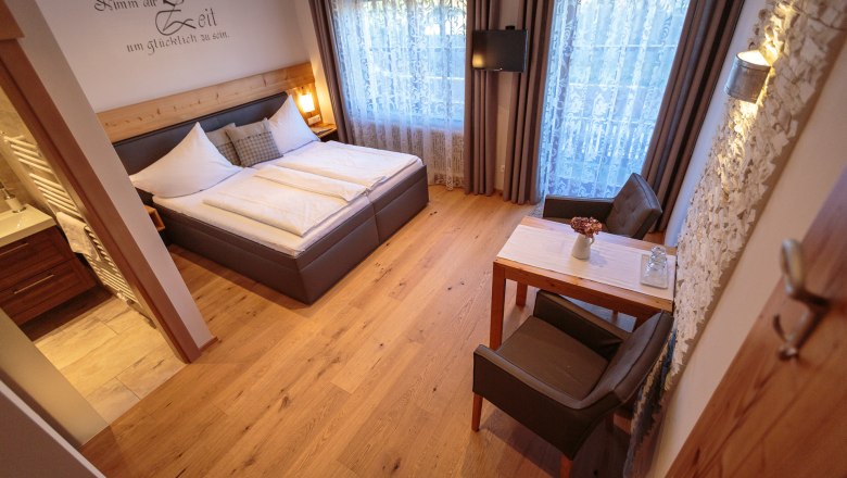 Útulná dvojlôžková izba s drevenou podlahou, manželskou posteľou, stolíkom a stoličkami, ozdobnými závesmi a TV na stene.