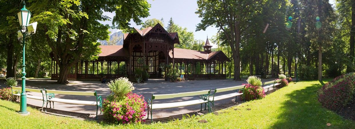 Pavilón v kúpeľných záhradách Reichenau, obklopený stromami a kvetinovými záhonmi.