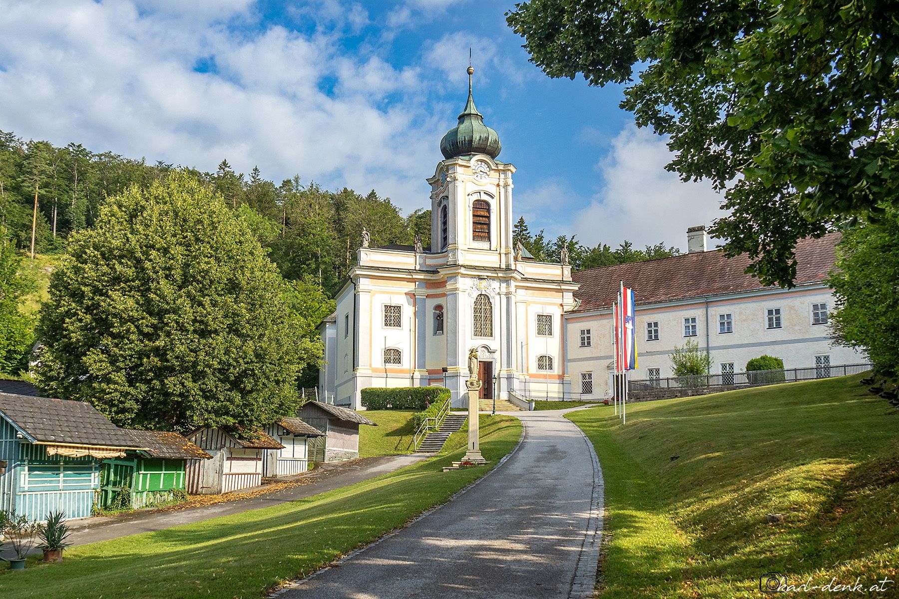 Servitský kláštor Mariahilfberg v Gutensteine s barokovým kostolom a vežou, obklopený stromami a cestou v popredí.