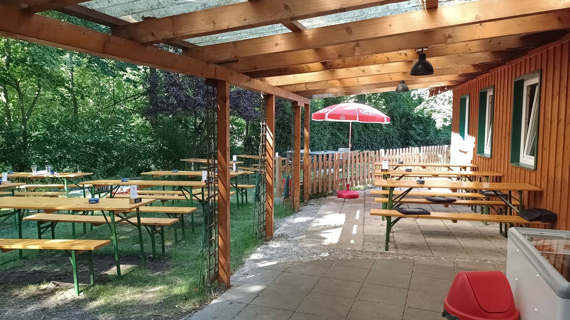 Krytá terasa s drevenými lavicami a stolmi, obklopená stromami a dreveným plotom.
