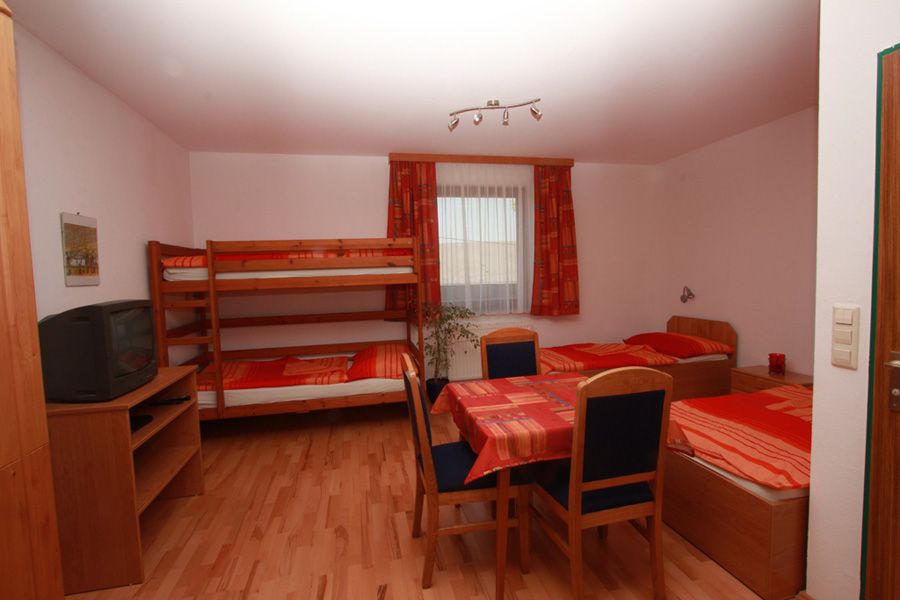 Jednoducho zariadená izba s poschodovou posteľou, samostatnou posteľou, stolom a stoličkami. Na poličke je umiestnená televízia. V pozadí je okno so závesmi.