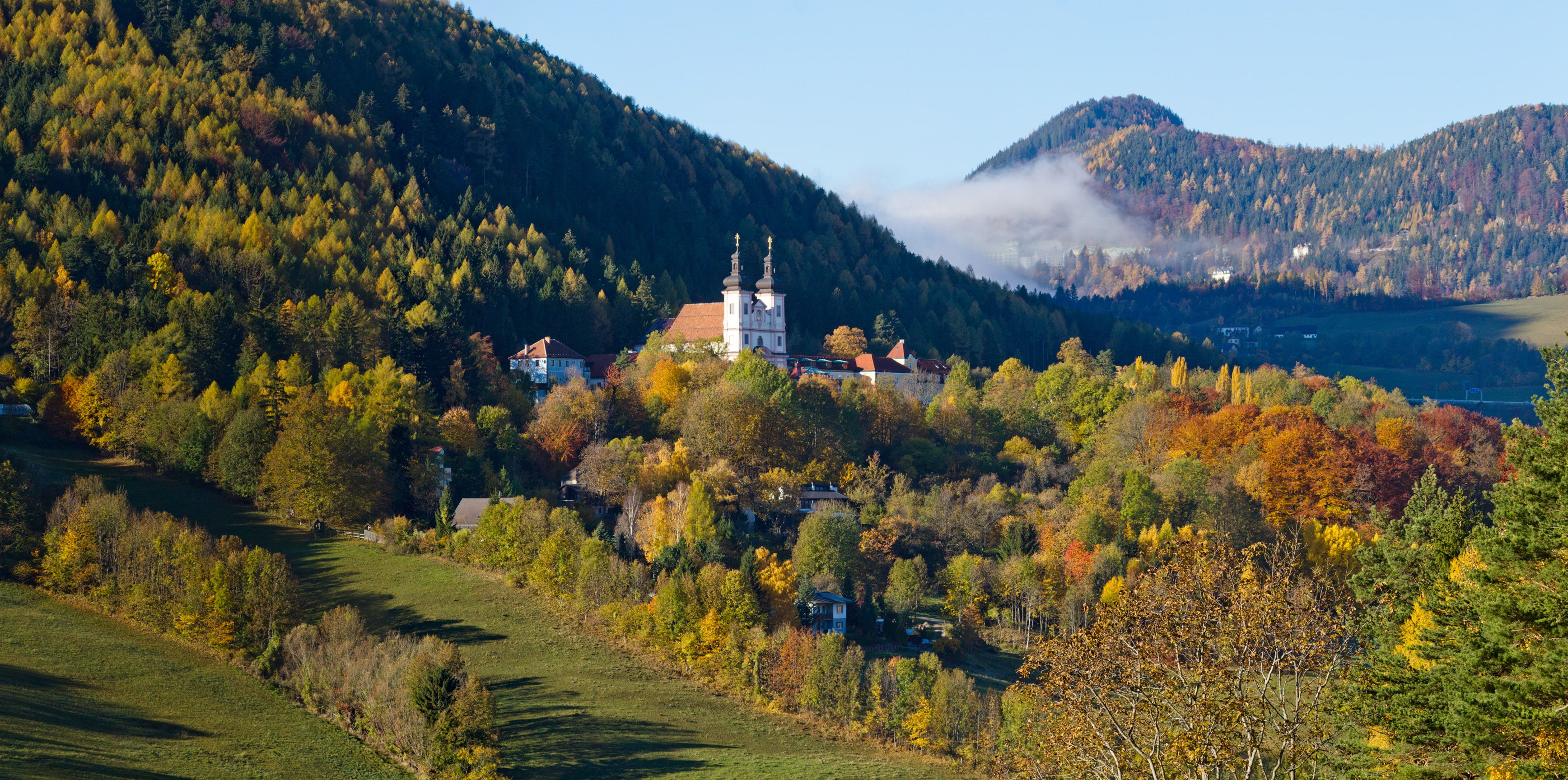 Jesenná krajina s kostolom v Maria Schutz, obklopená farebnými stromami a kopcami.