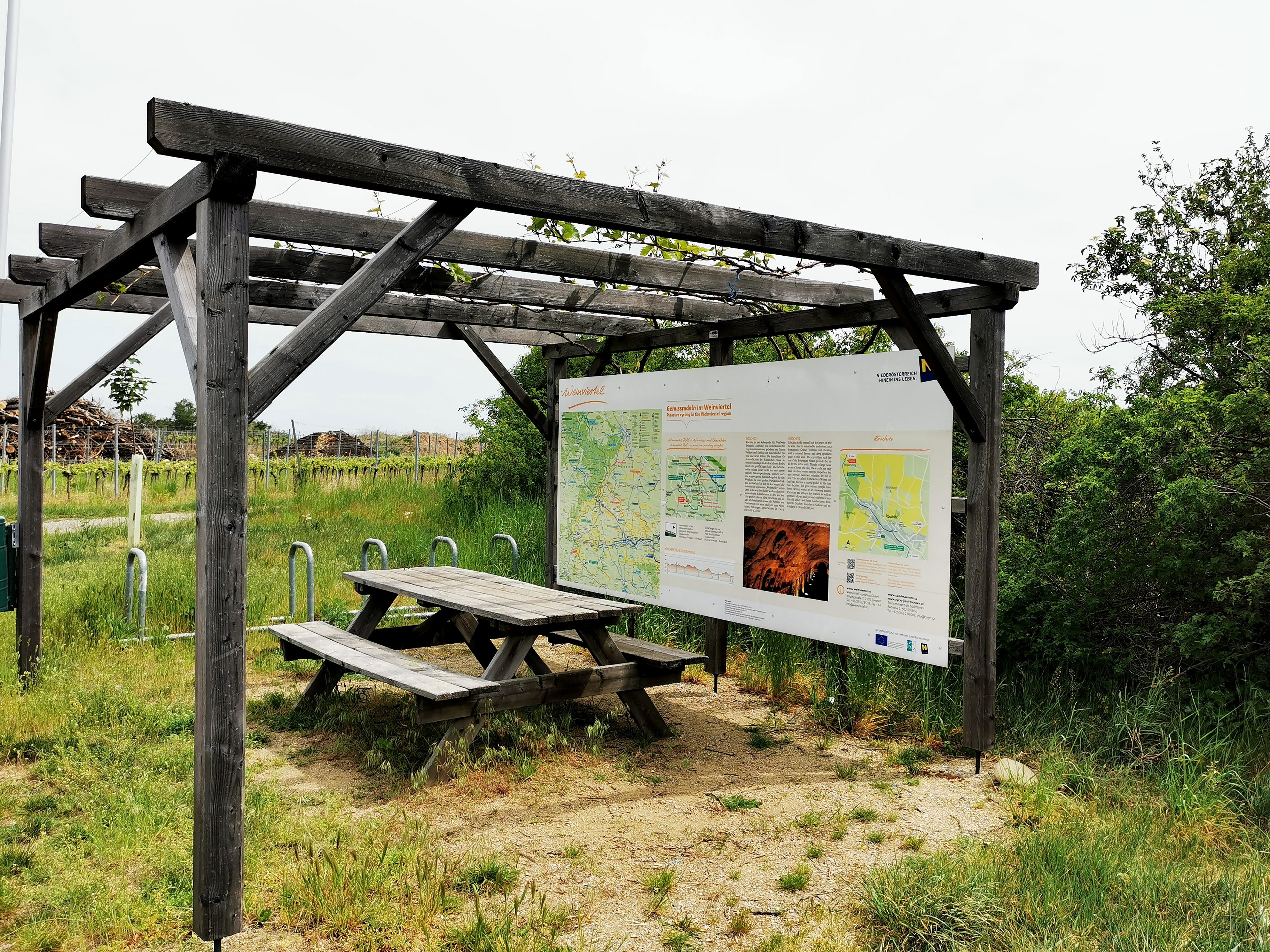 Drevená pergola s informačnou tabuľou a piknikovým stolom vo vidieckom prostredí.