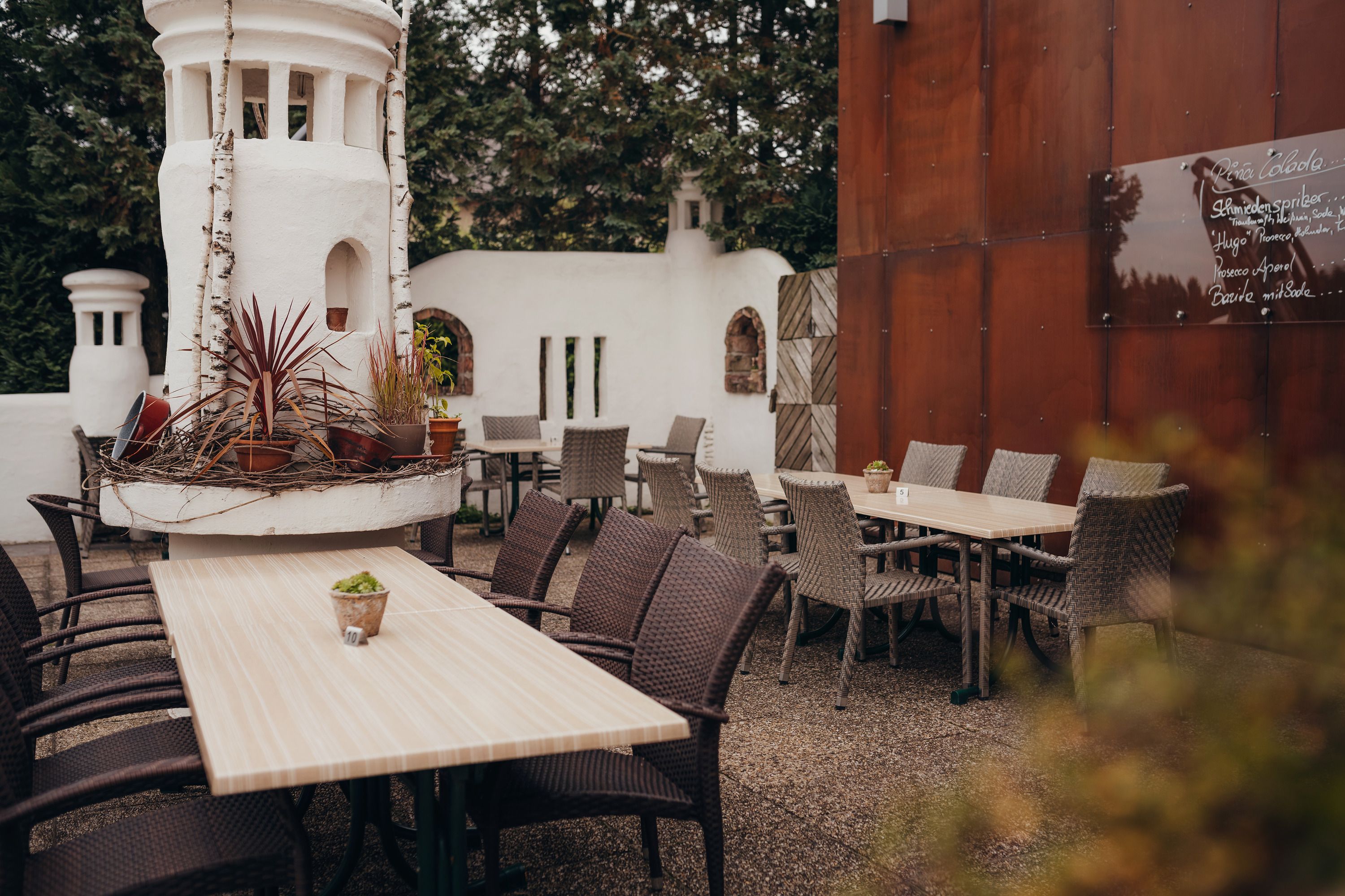 Útulná záhrada pre hostí s drevenými stolmi, tkanými stoličkami a okrasnými rastlinami v bielej veži.