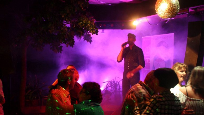 Spev&aacute;k na p&oacute;diu s mikrof&oacute;nom, obklopen&yacute; tancuj&uacute;cimi p&aacute;rmi a farebn&yacute;mi svetlami.