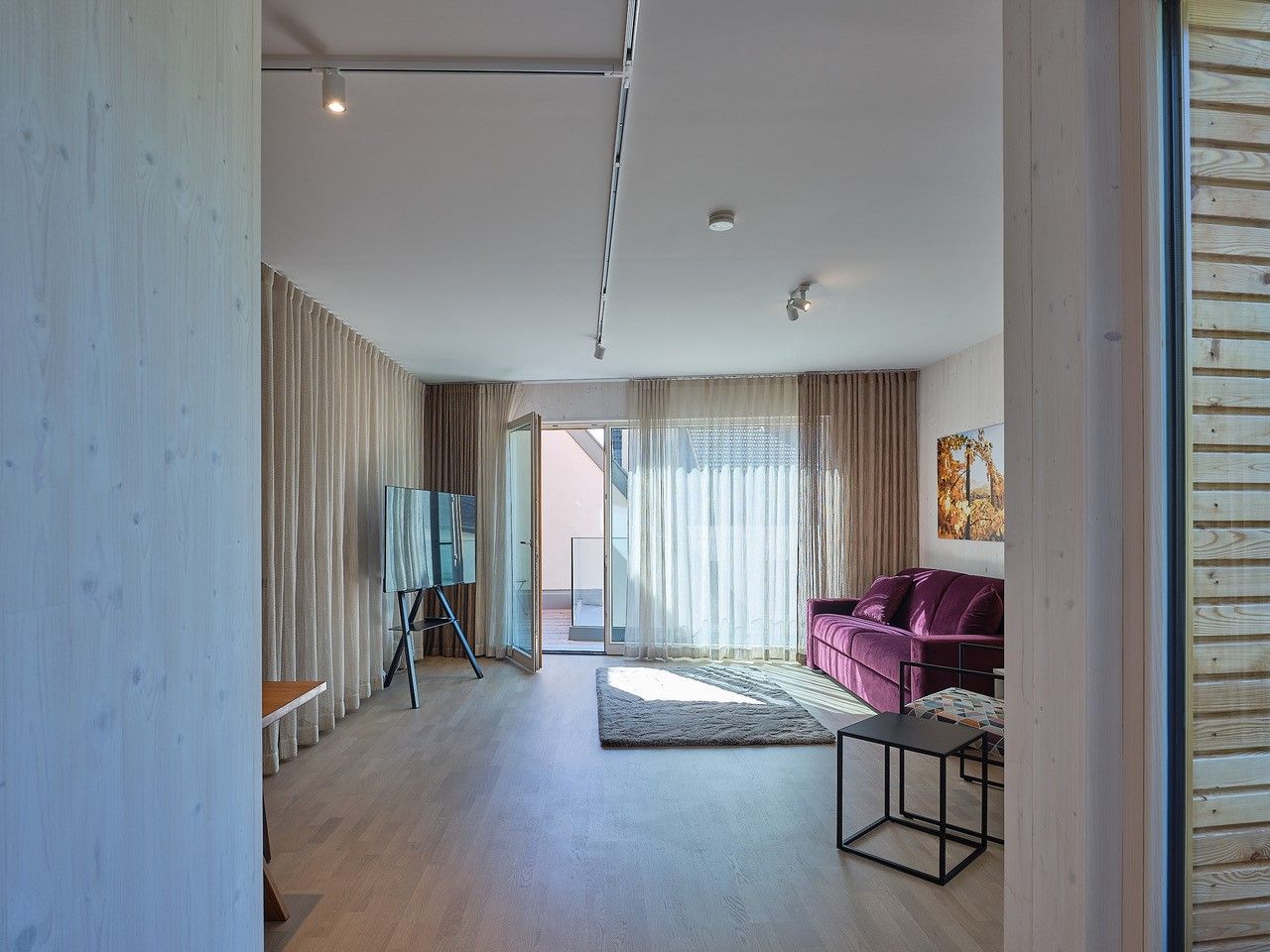 Moderná obývacia izba s fialovou pohovkou, veľkým oknom a TV na stojane.