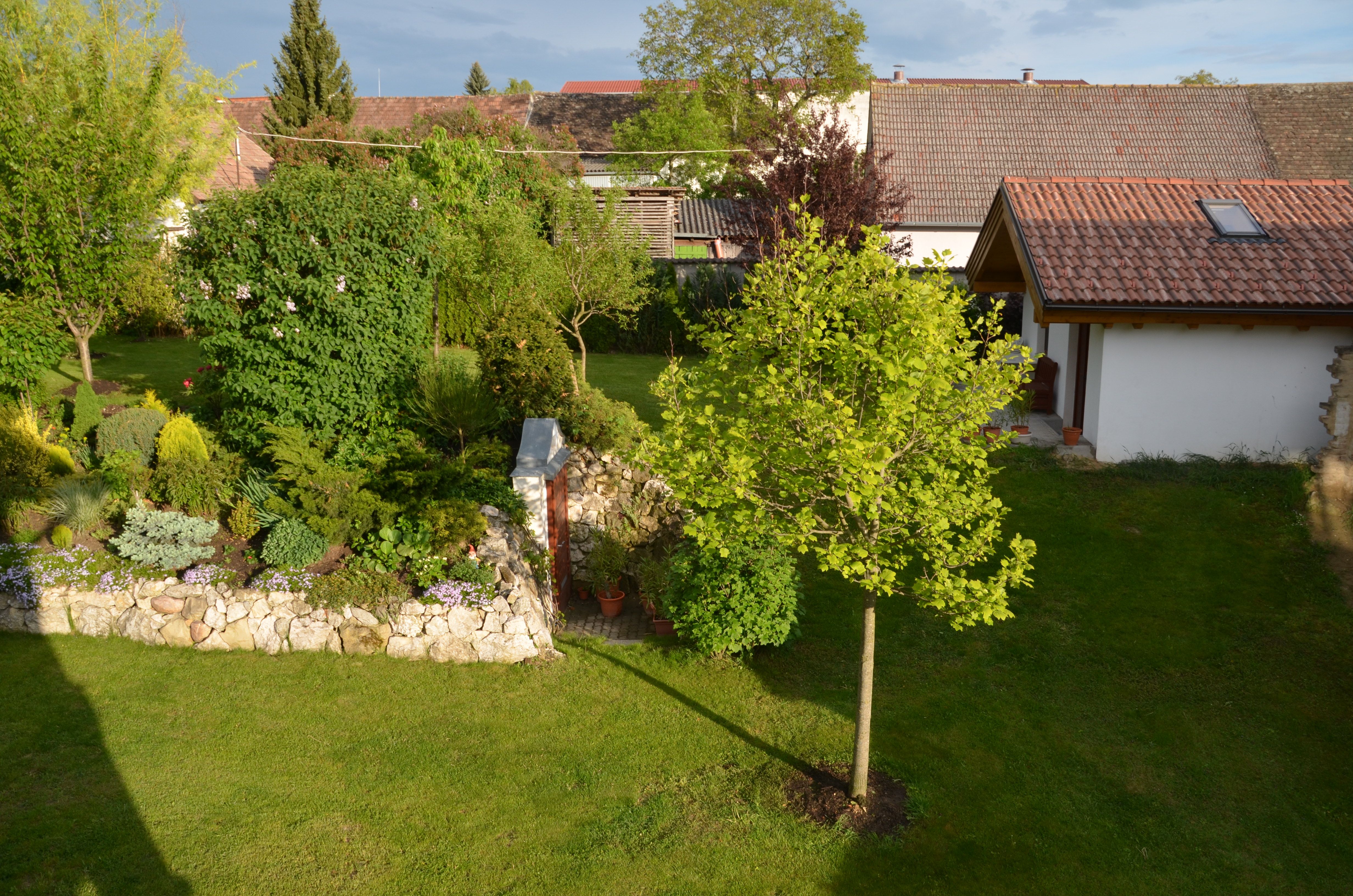 Udržiavaná záhrada so stromami, kríkmi a malou budovou so škridlovou strechou.