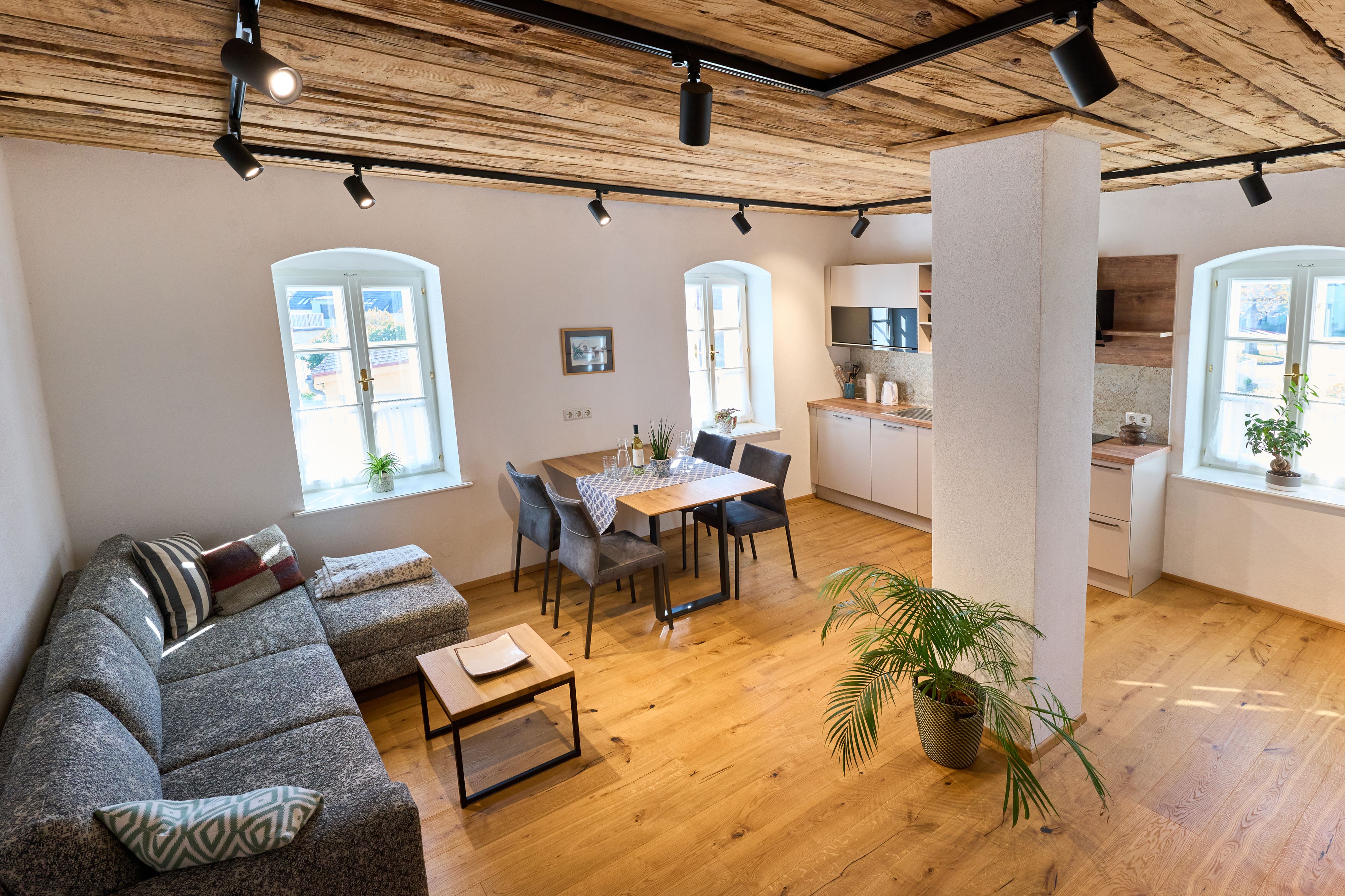 Moderná obývacia izba s drevenou podlahou, sivou pohovkou, jedálenským stolom a kuchyňou.