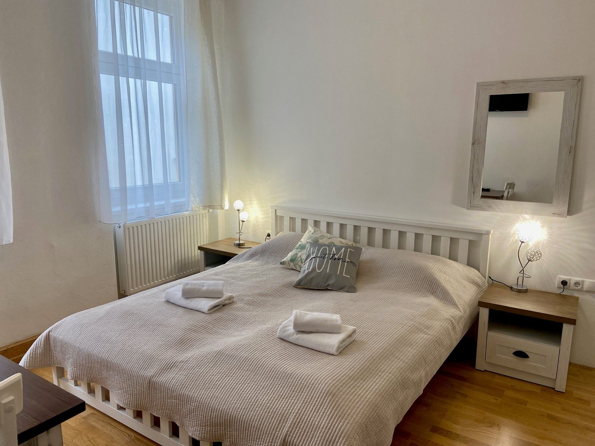 Útulná dvojlôžková izba s veľkou posteľou, dvoma nočnými stolíkmi a zrkadlom na stene. Izba je svetlá a príjemná.