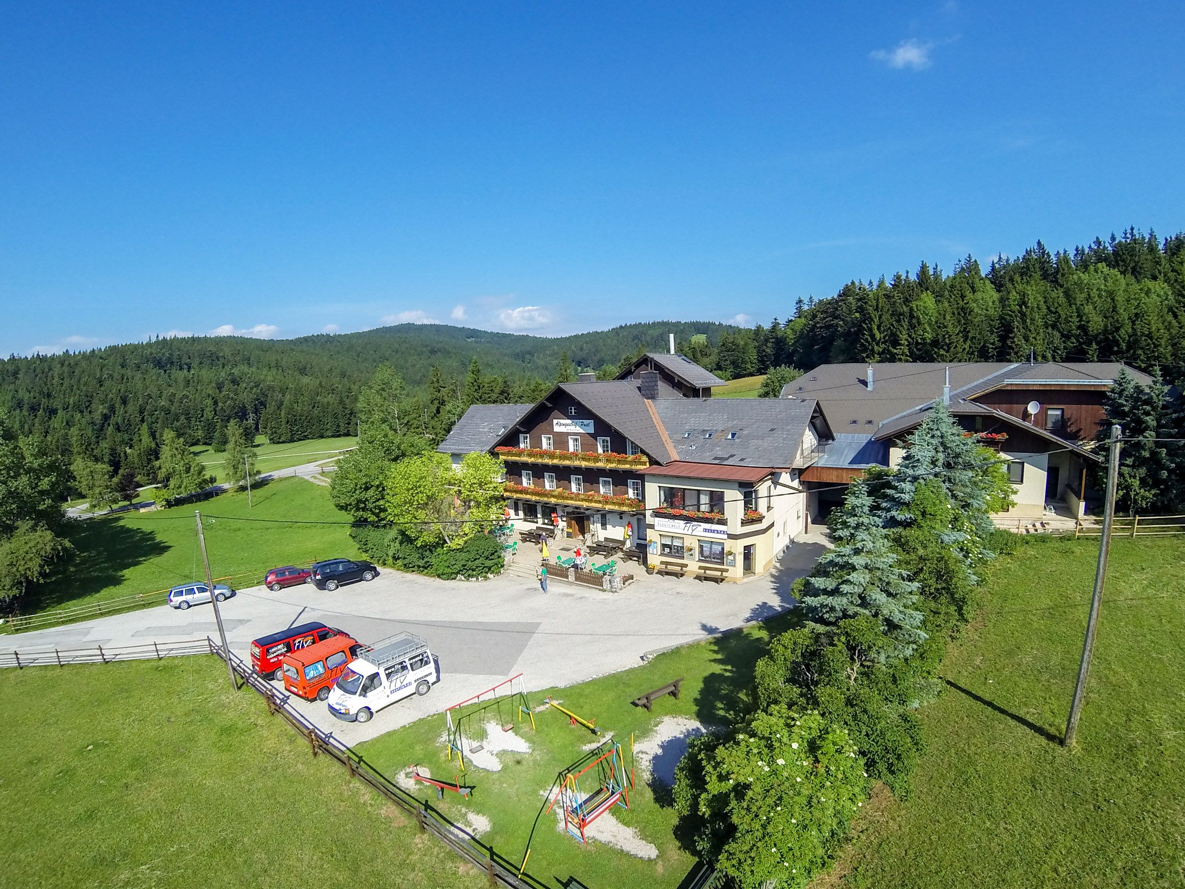 Alpengasthof Postl v zelenej zalesnenej krajine s niekoľkými zaparkovanými autami a detským ihriskom v popredí.