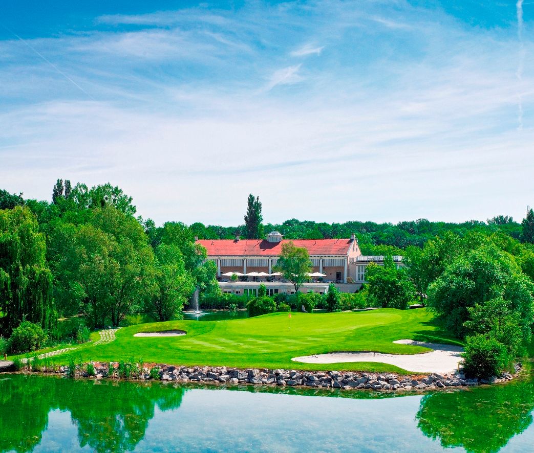 Golfové ihrisko s klubovňou v pozadí, obklopené stromami a rybníkom.