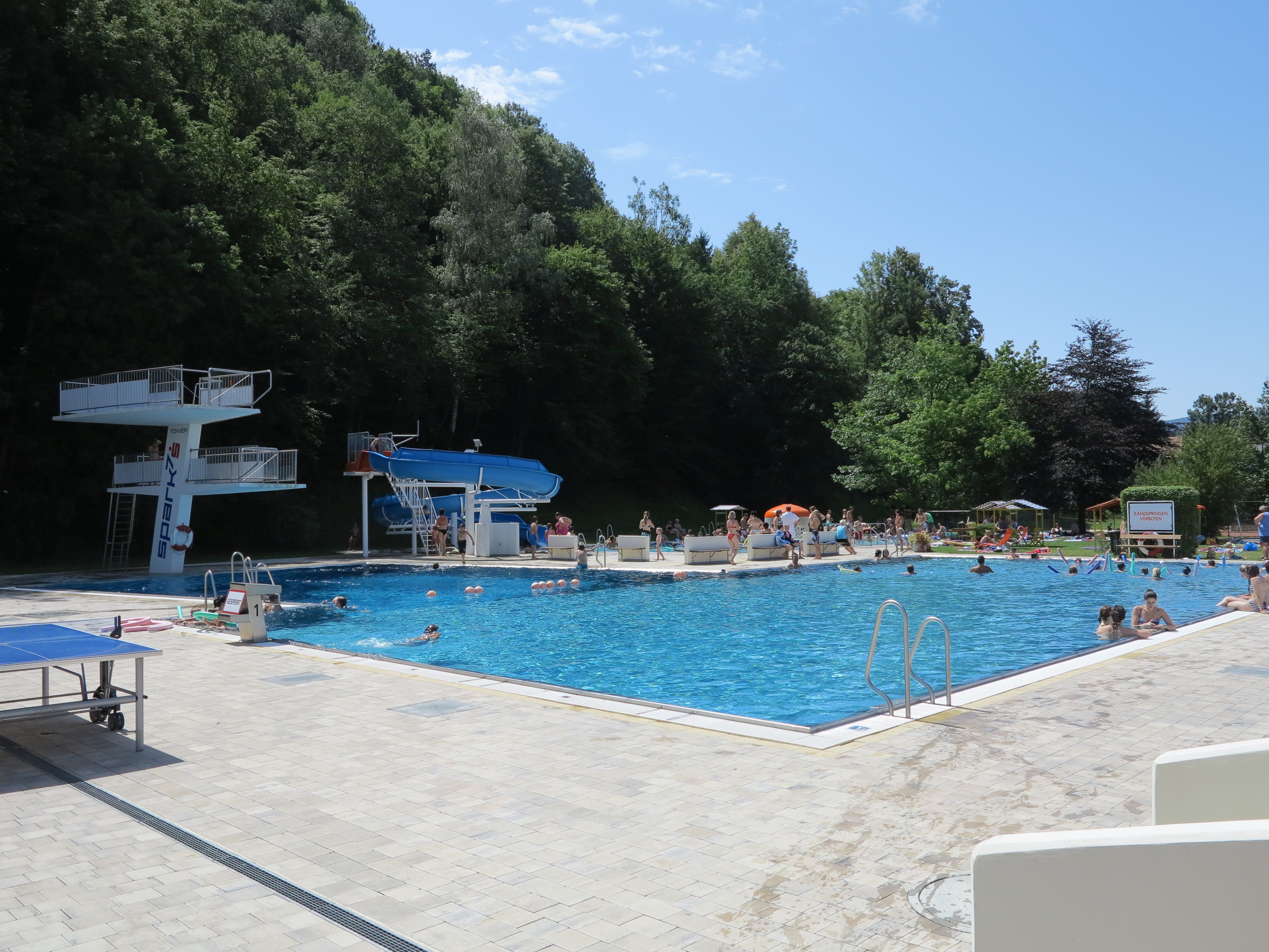Vonkajší bazén so skokanskou plošinou, šmykľavkou a množstvom kúpajúcich sa počas slnečného dňa.