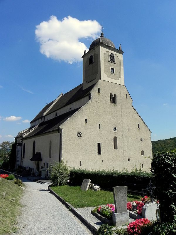 Historický kostol s vežou a okolitým cintorínom pod jasnou oblohou.