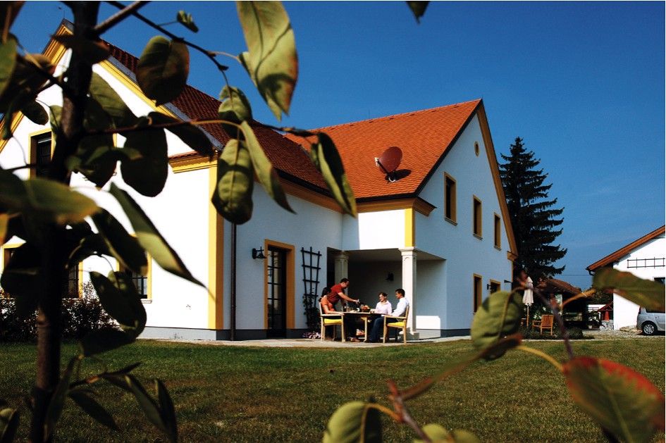 Biely dom s červenou strechou, obklopený zeleným trávnikom a stromami. Ľudia sedia vonku pri stole.