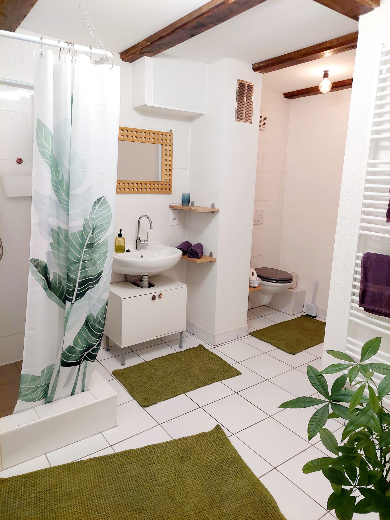 Moderná kúpeľňa s bielymi obkladačkami, zelenými kobercami a sprchovým závesom s rastlinným motívom.