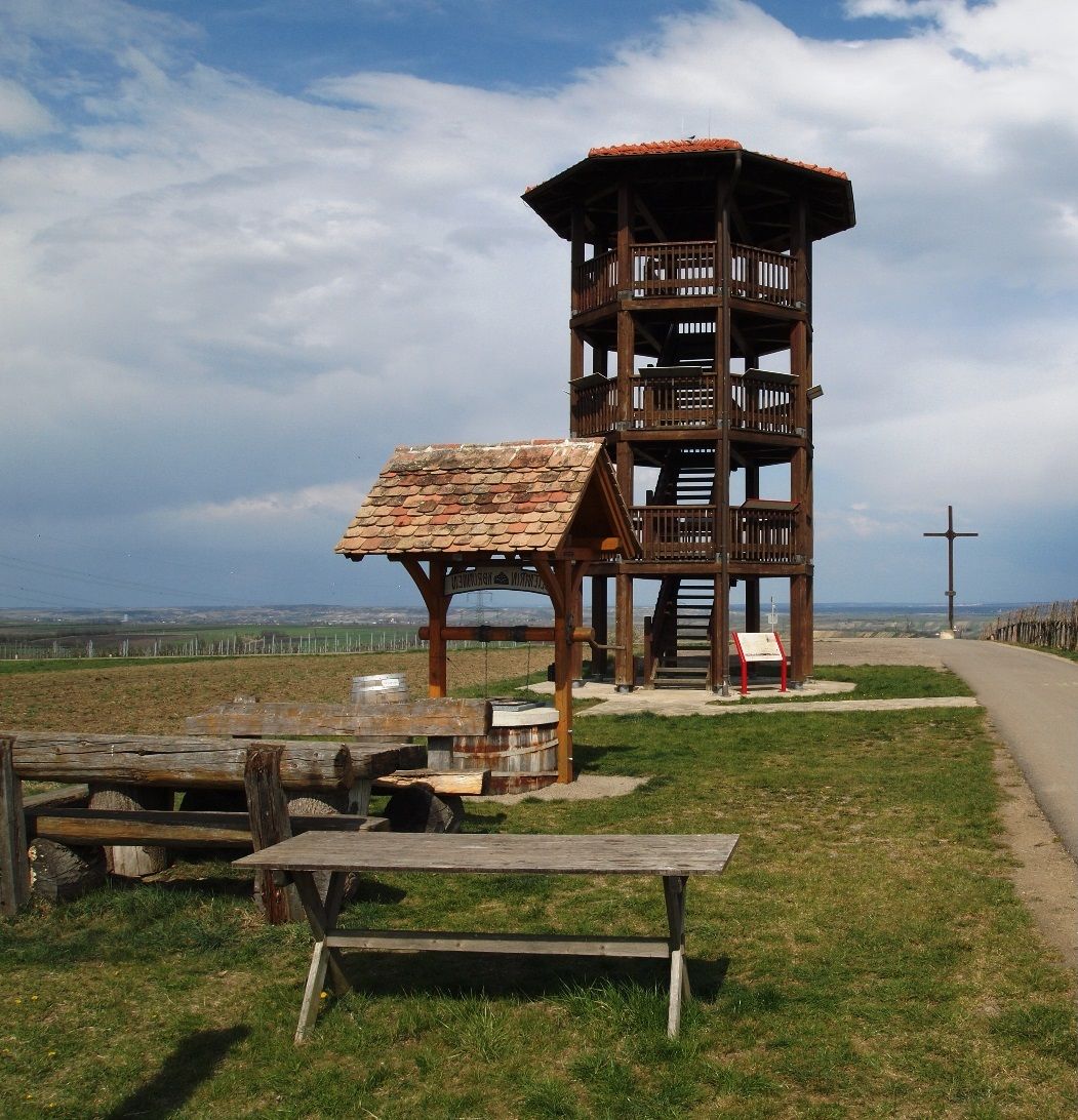 Drevená rozhľadňa na lúke s lavičkami a krížom v pozadí.