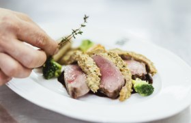 Detailný záber na ružové pečené mäso s bylinkovou kôrkou, brokolicou a rukou pridávajúcou tymián.