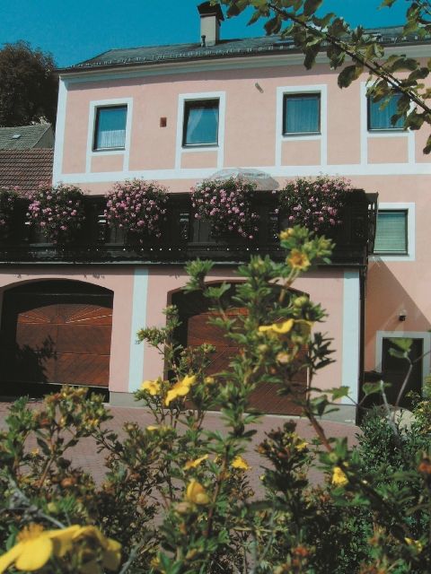 Dvojposchodová ružová budova s kvitnúcimi rastlinami na balkóne a žltými kvetmi v popredí.