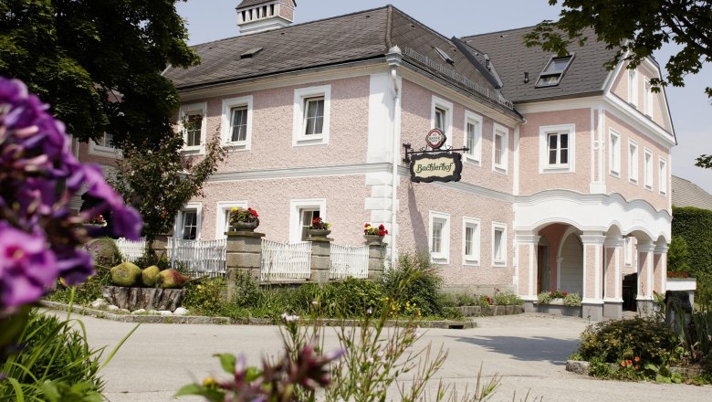 Tradičná, ružovo natretá budova s nápisom "Bachlerhof", obklopená kvetmi a stromami.