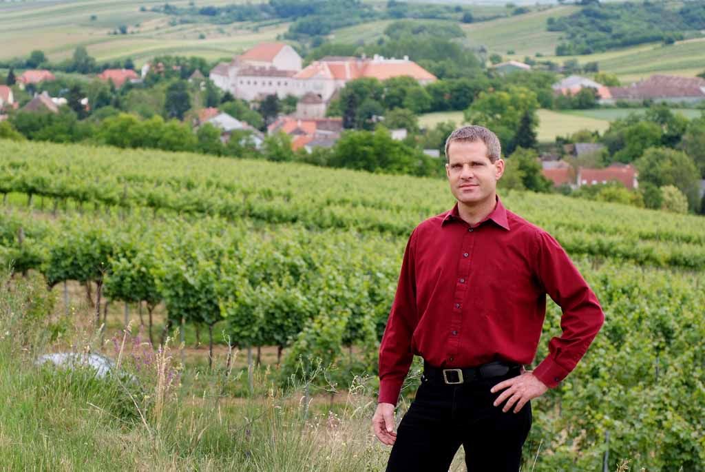 Osoba v červenej košeli stojí pred vinicami s dedinou v pozadí.