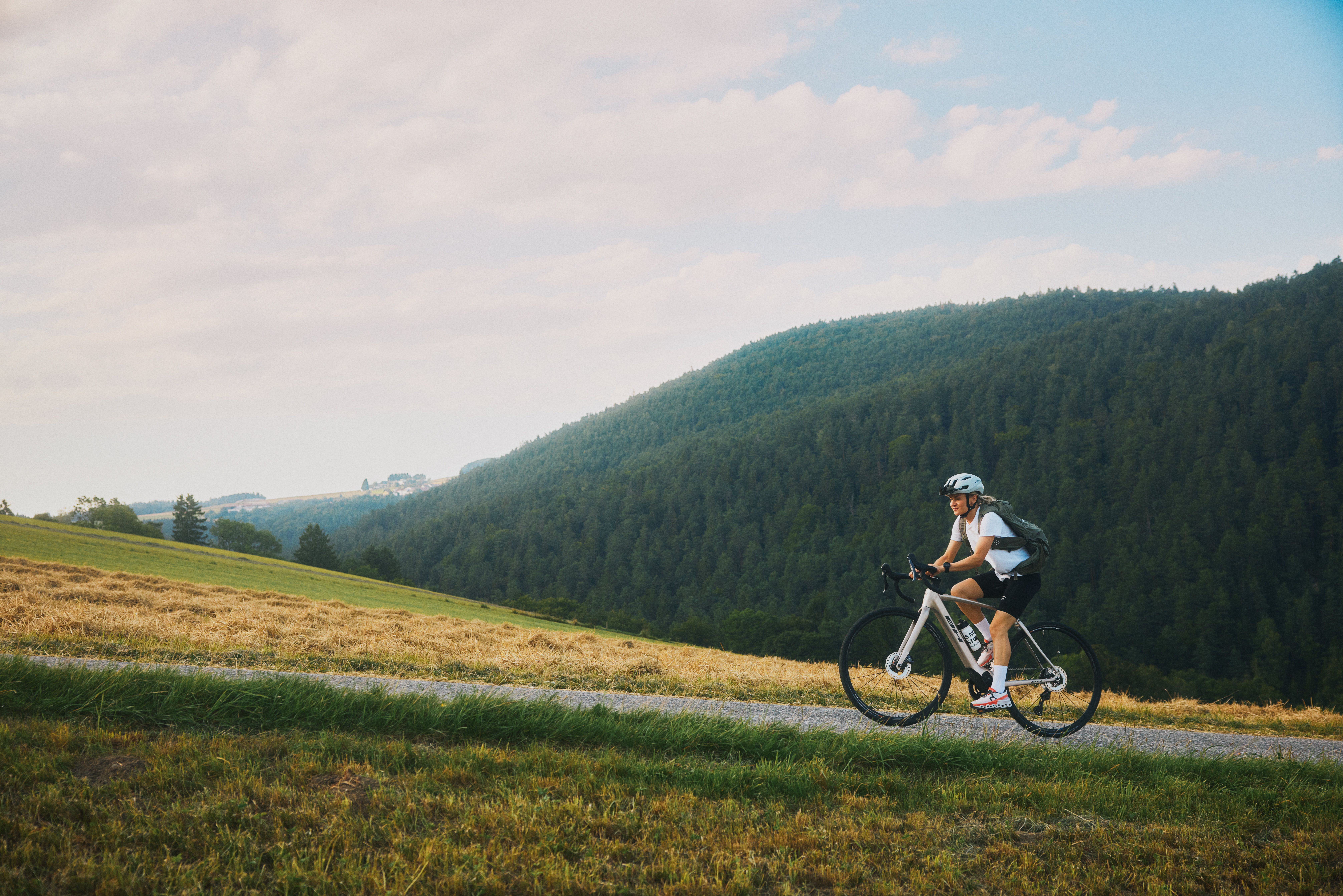 Cyklista jazdí po úzkej stúpajúcej ceste so zalesnenými kopcami v pozadí.
