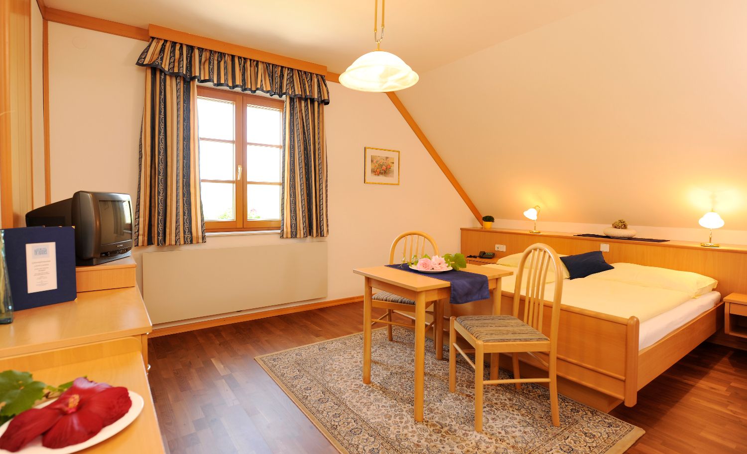 Útulná izba pre hostí s dreveným nábytkom, manželskou posteľou, stolíkom a stoličkami, TV a oknom so závesmi.