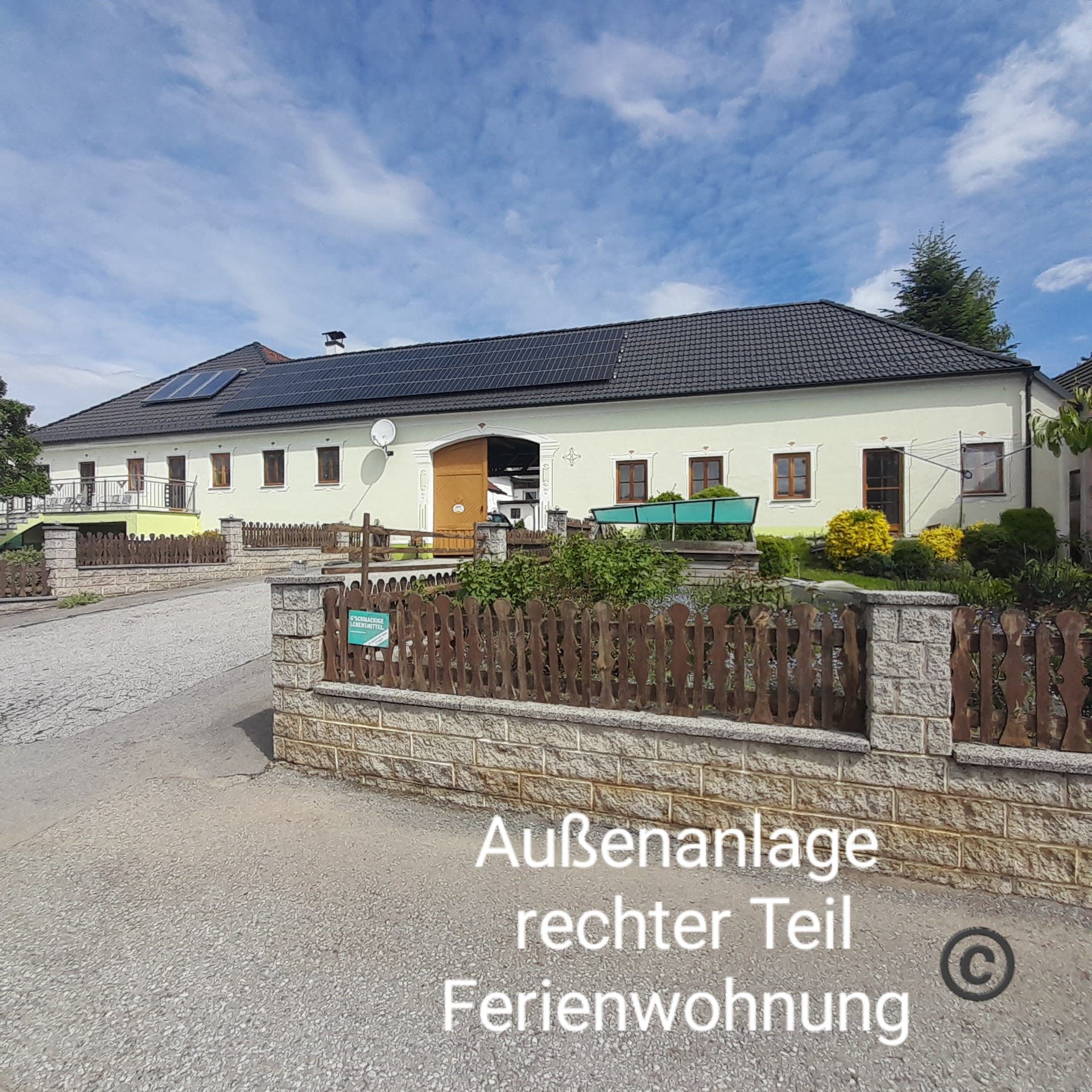 Veľká budova so žltou fasádou a čiernou strechou, obklopená záhradou a dreveným plotom. Obloha je modrá s niekoľkými mrakmi.