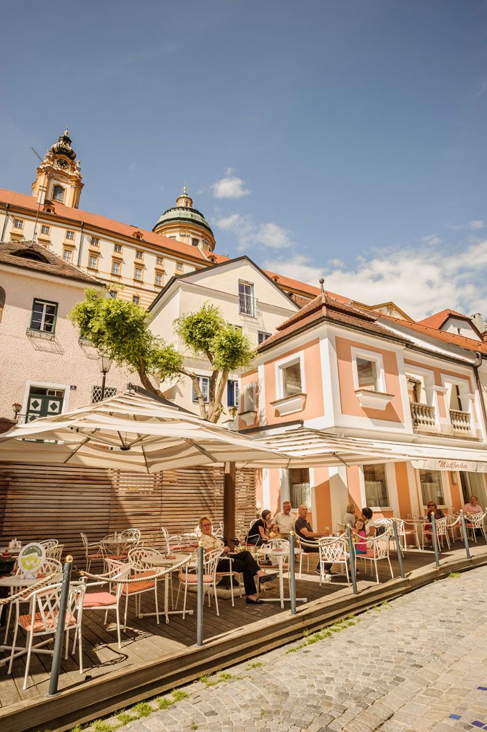 Vonkajší priestor kaviarne so slnečníkmi a hosťami, v pozadí historická budova s kupolou.