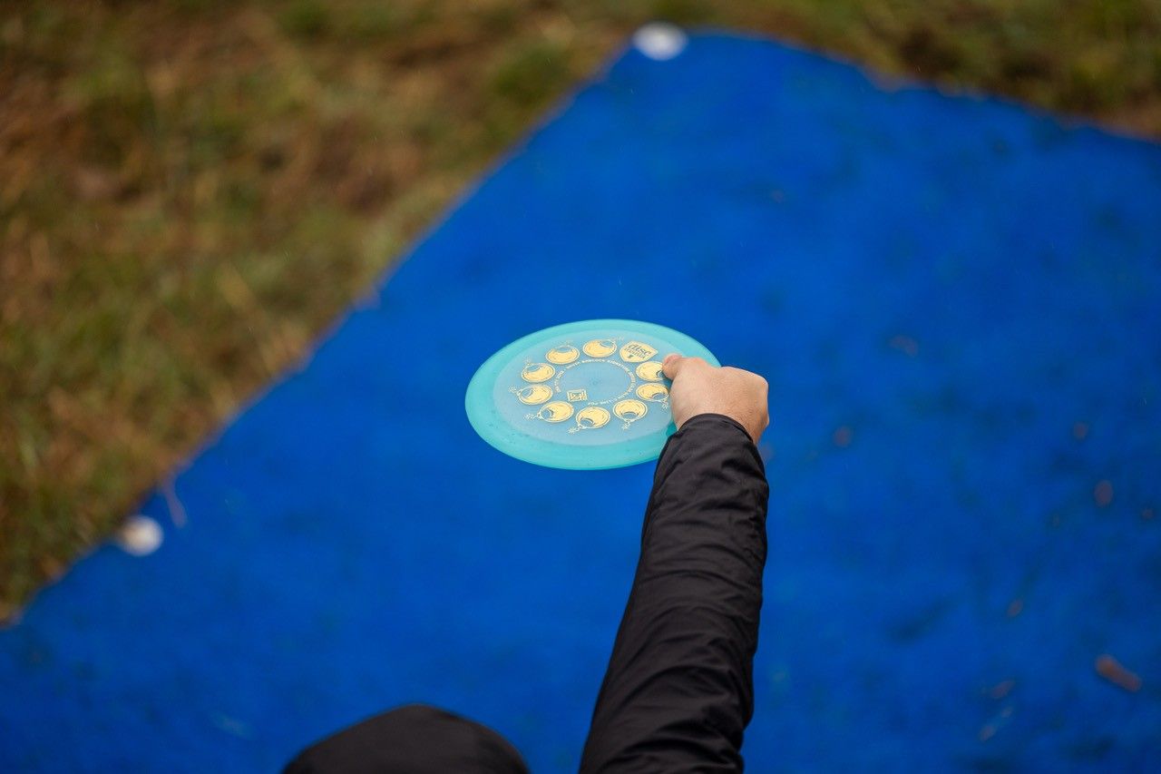 Osoba drží discgolfový disk nad modrou podložkou, ruku natiahnutú na hod