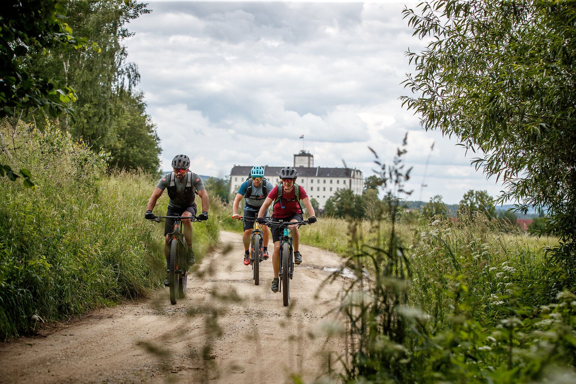 Traja horskí cyklisti sa kĺžu po úzkej prašnej ceste obklopenej sviežou zeleňou a kopcami. Svieži horský vzduch a úchvatná scenéria vás pozývajú na nezabudnuteľné dobrodružstvo. Tu, kde sa stretáva príroda a šport, sa každý okamih stáva zážitkom.