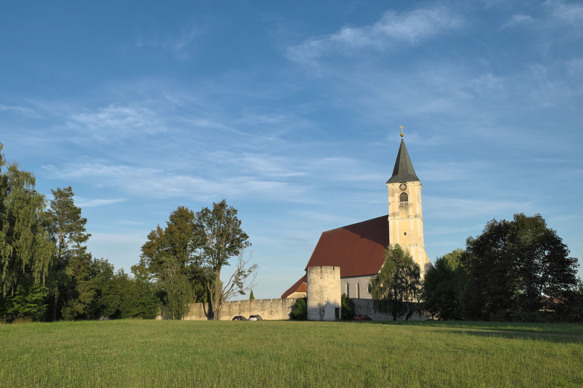 Kostol s vežou a červenou strechou na lúke, obklopený stromami a múrom.