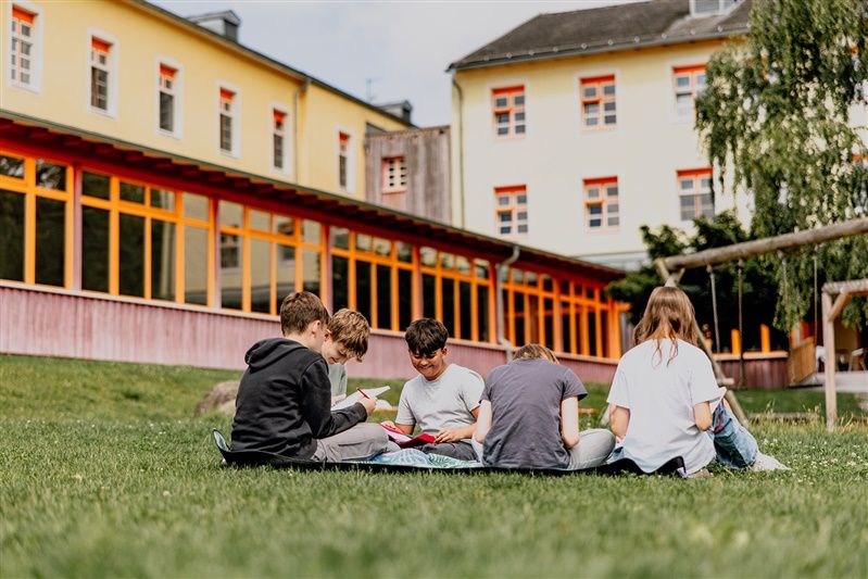 Skupina detí sedí na lúke pred žltou budovou.