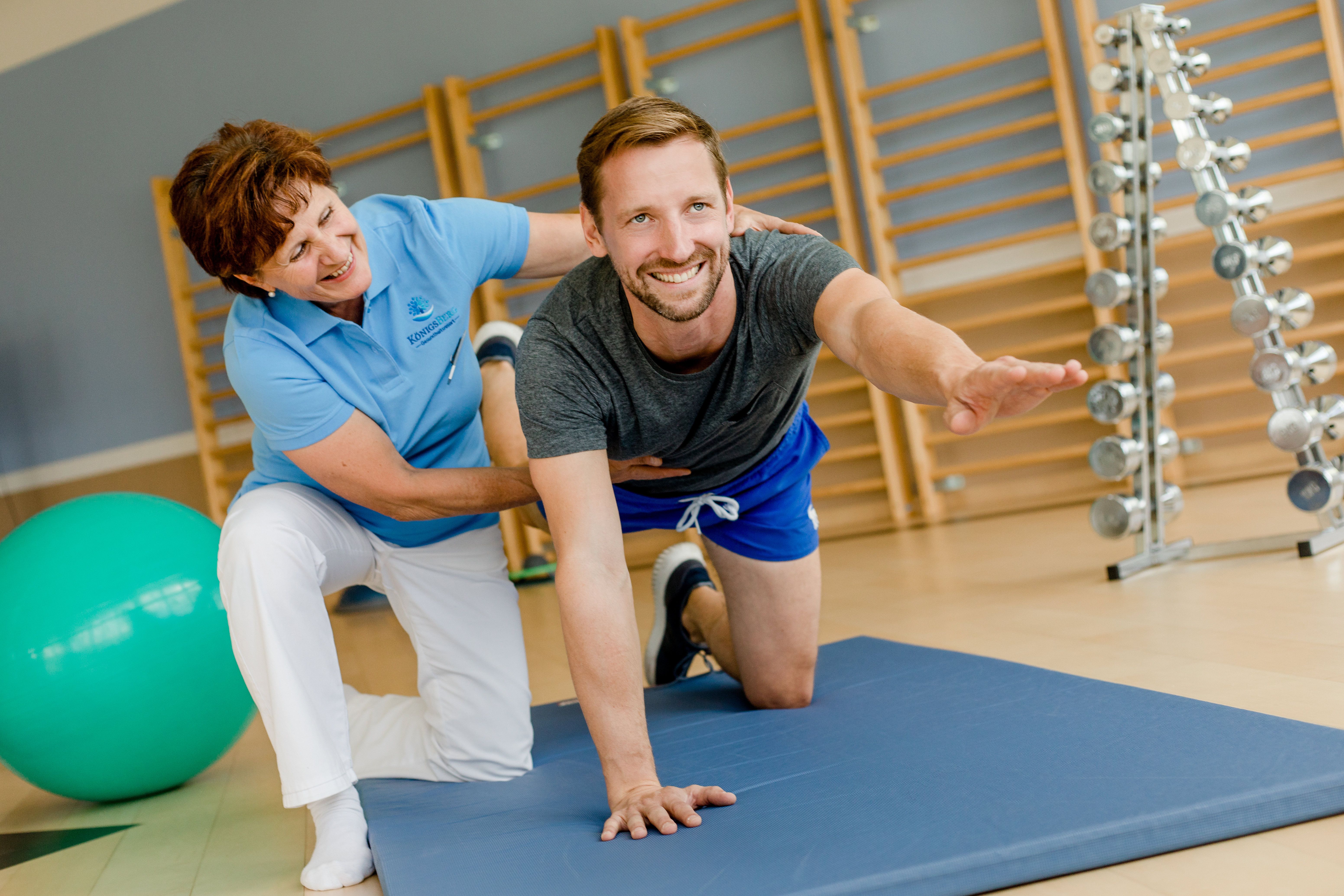 Muž cvičí na podložke s podporou terapeuta vo fitnescentre.