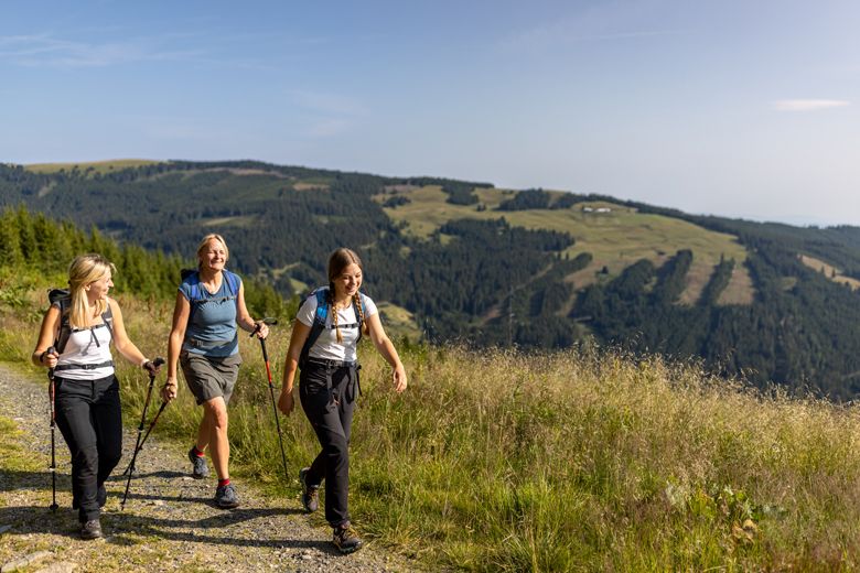 Tri ženy kráčajú po horskej ceste s turistickými palicami a ruksakmi, obklopené zelenými kopcami a lesmi.